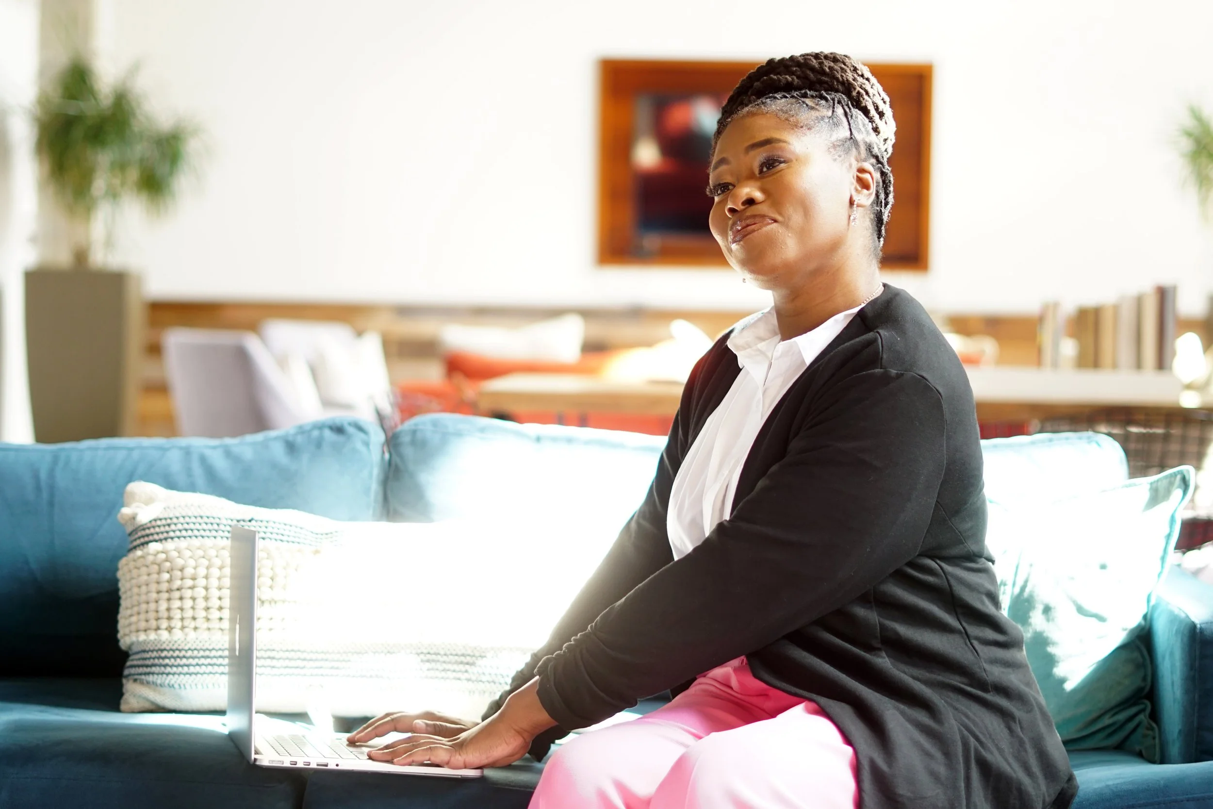A woman sitting on a blue couch in a bright room, using a laptop, with abstract artwork and a bookshelf in the background.