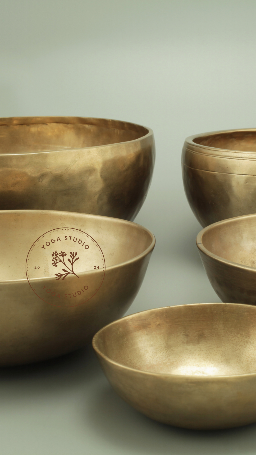 A collection of five bronze singing bowls of various sizes arranged on a surface with a light, neutral background. The bowls are metallic and reflective.
