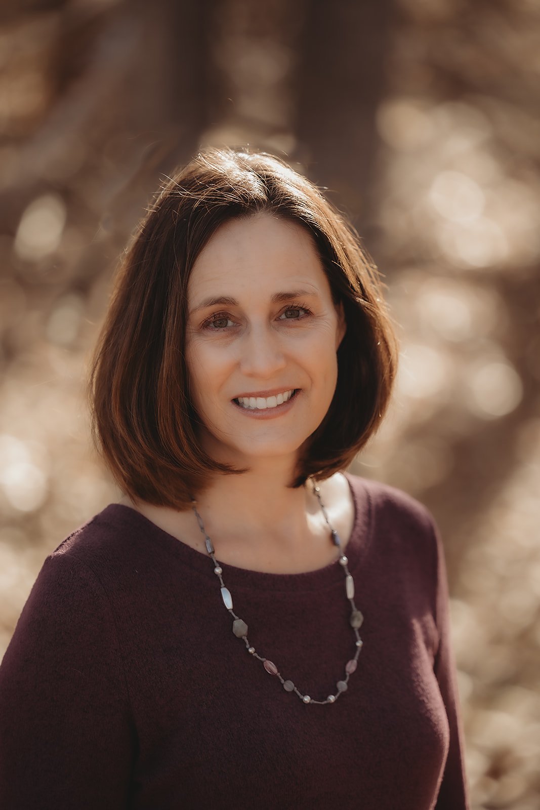 A woman with shoulder-length brown hair, wearing a maroon top and a beaded necklace, standing outdoors with a blurred background of trees and sunlight.