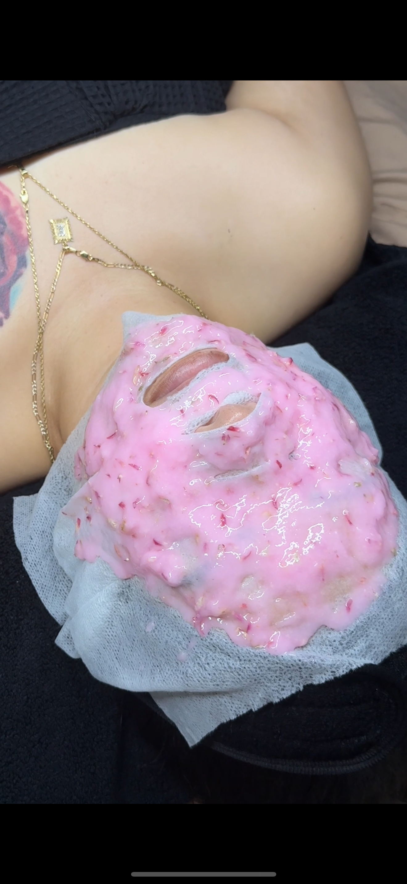 A person lying down with a pink facial mask applied to their face, wearing a gold chain necklace, and a black top, with part of a tattoo visible on their shoulder.