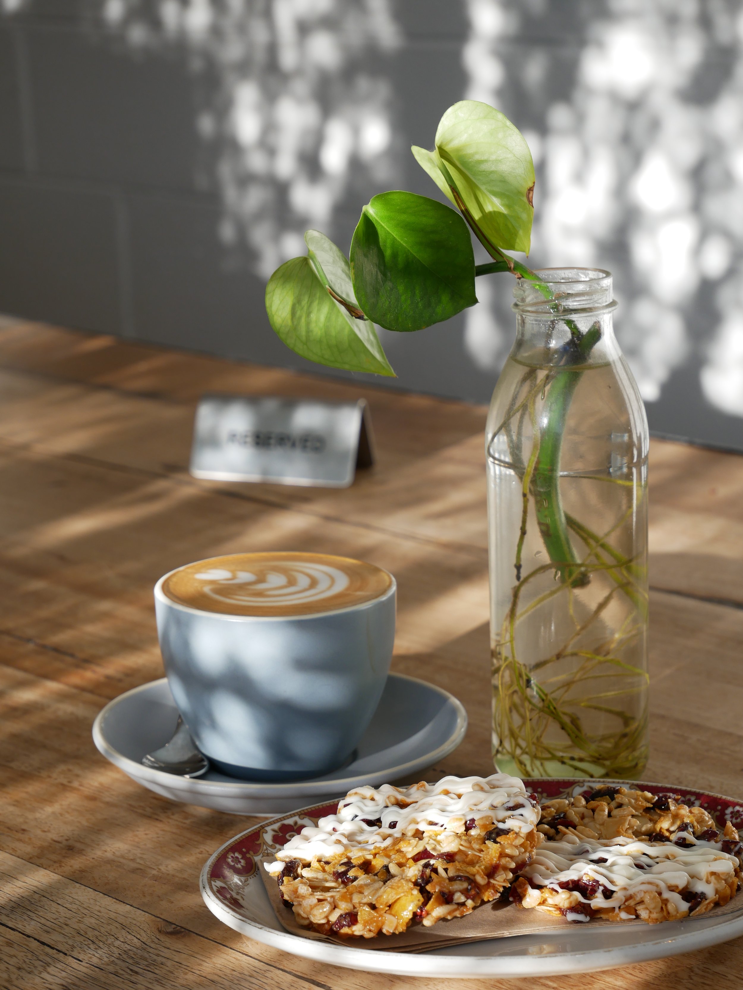 A table with a latte in a blue cup on a saucer, two nut and dried fruit bars with icing on top on a plate, a glass vase with green leaves, and blurred background.