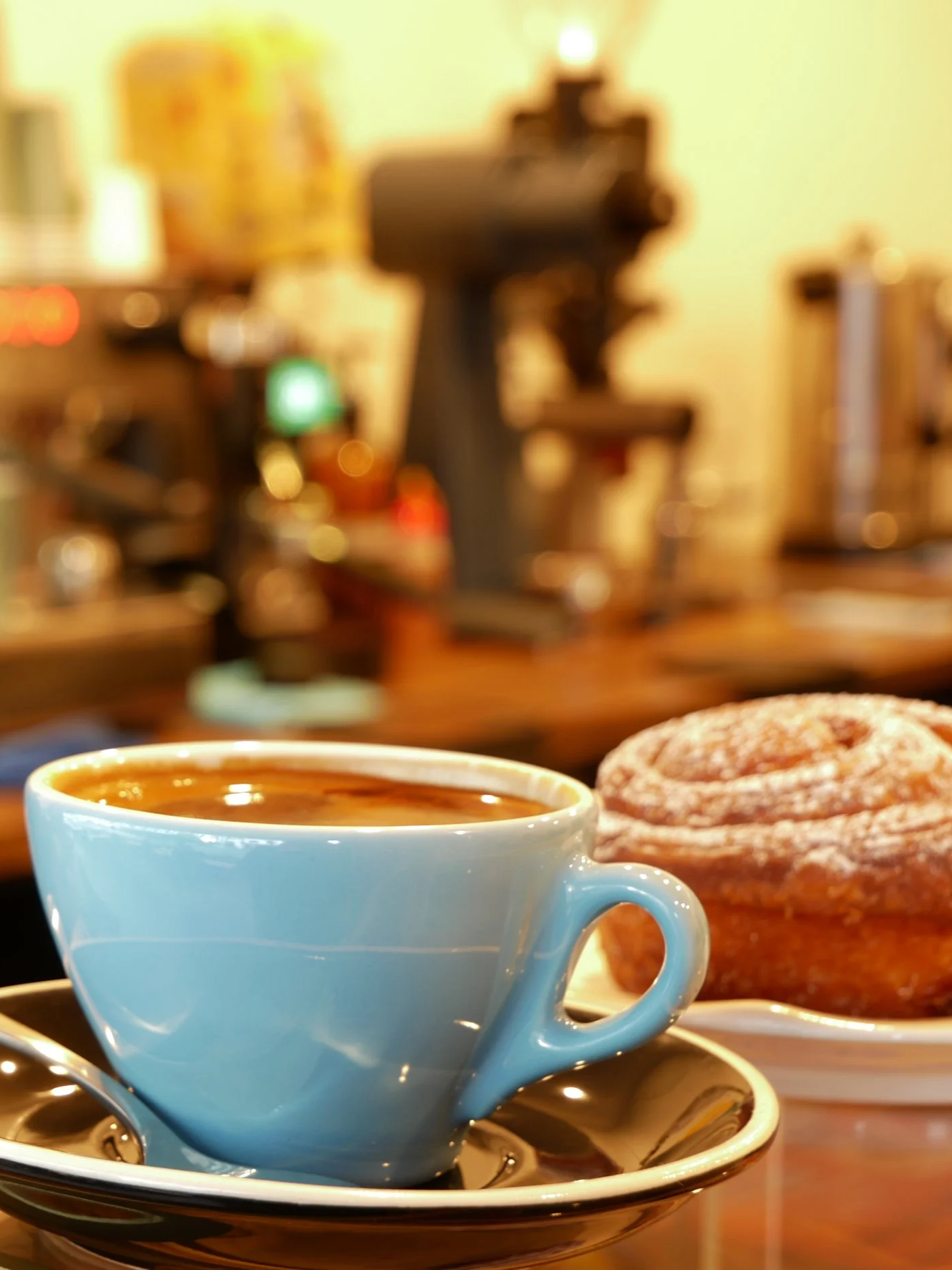 A blue coffee cup filled with coffee on a saucer, with sugar-coated donuts on a plate in the background, in a cozy cafe setting.