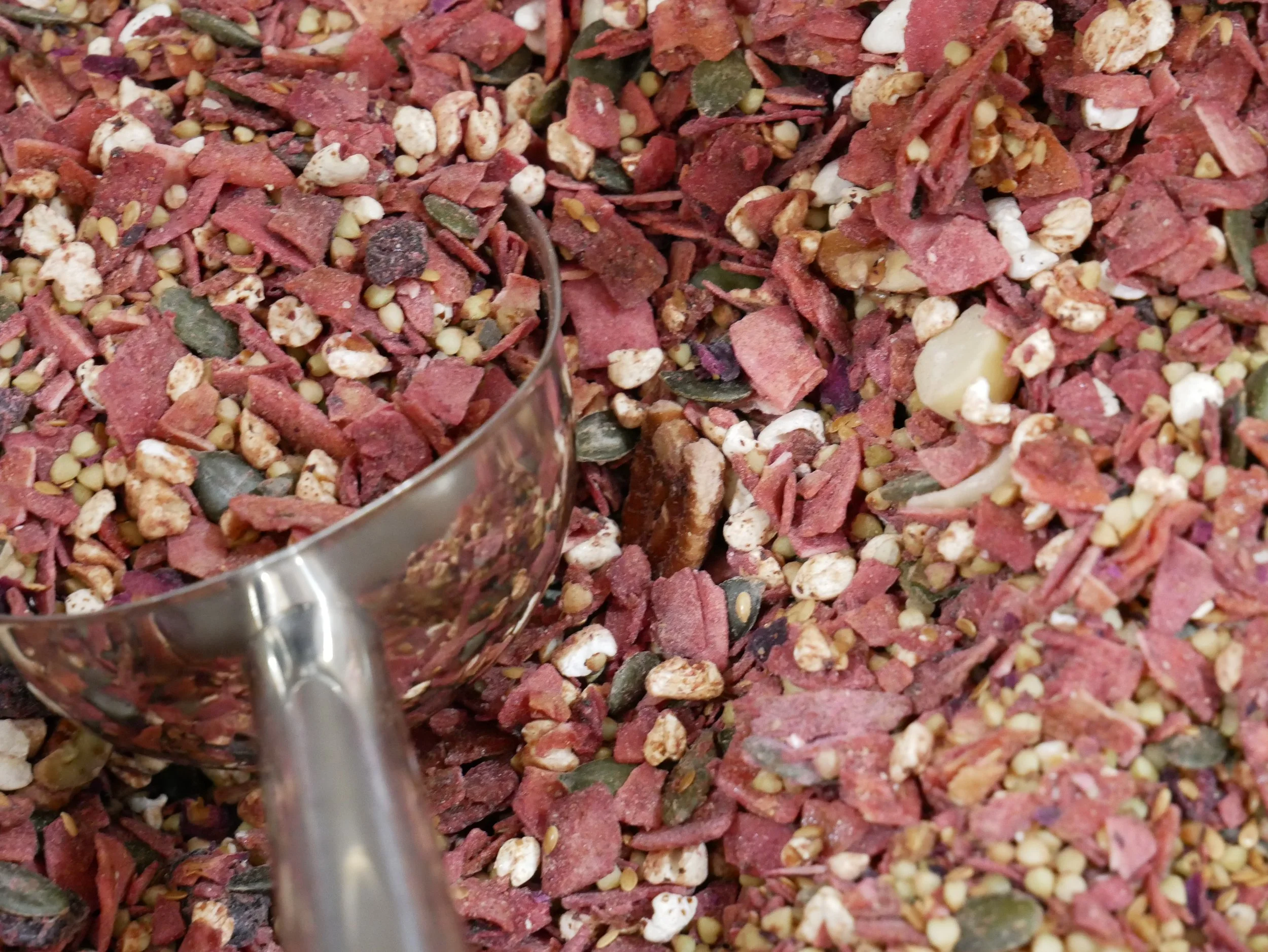 Close-up of a mixture of dried herbs and spices, including red flakes, green leaves, and small beige seeds, with a metal spoon in the foreground.