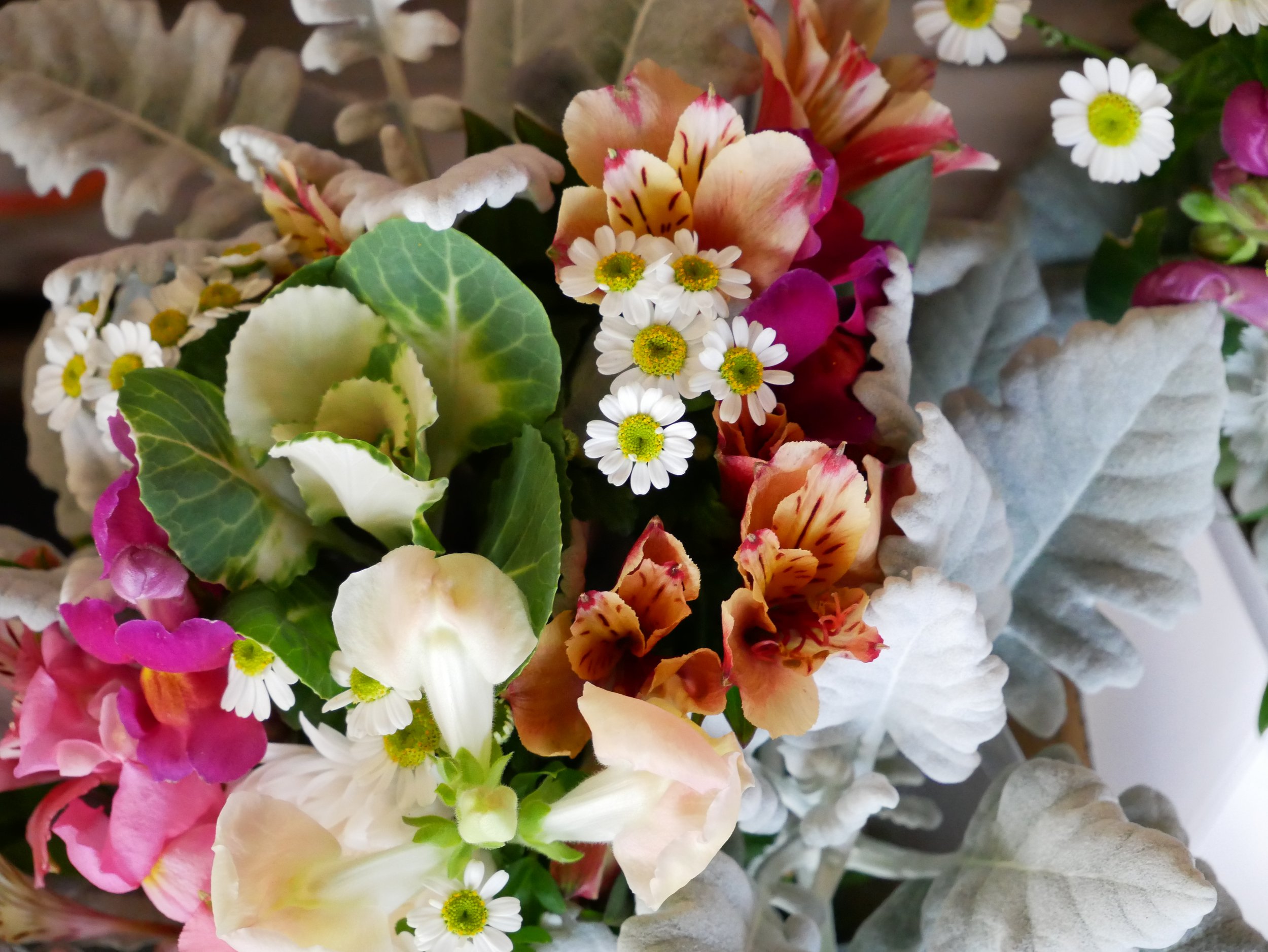 Close-up of a bouquet of various colorful flowers including daisies, roses, and other blossoms with different shades of pink, white, orange, and yellow, and green leaves.