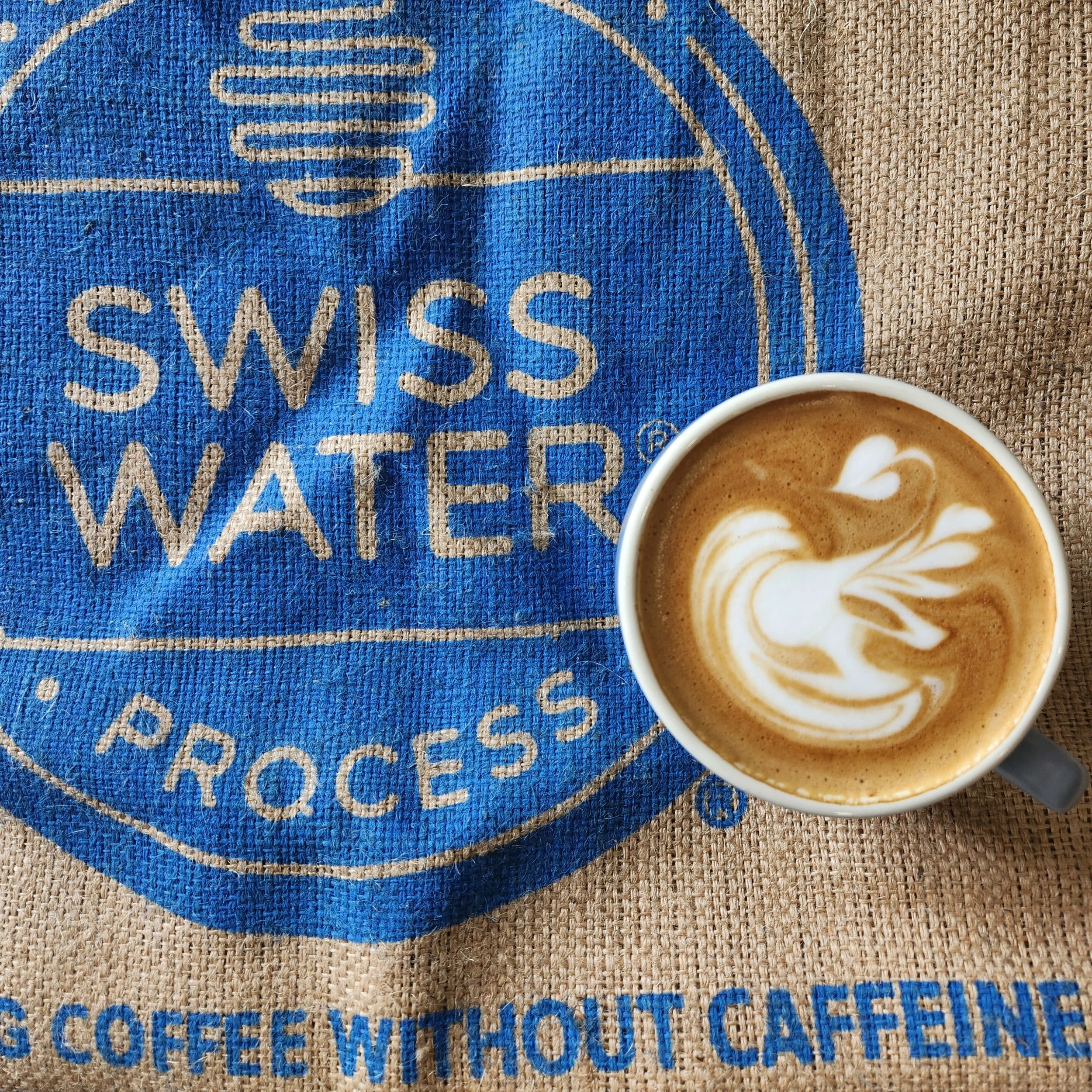 Close-up of a coffee cup with latte art, placed on a woven beige surface. Part of a blue cloth with stitched design and text is visible underneath the cup.