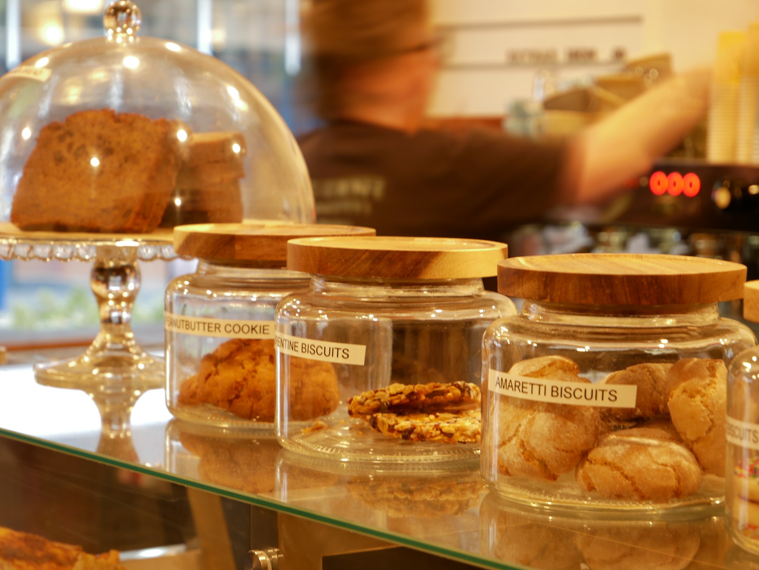 Display of glass jars containing various types of cookies, including peanut butter and amaretti biscuits, with a cake under a glass dome in the background.