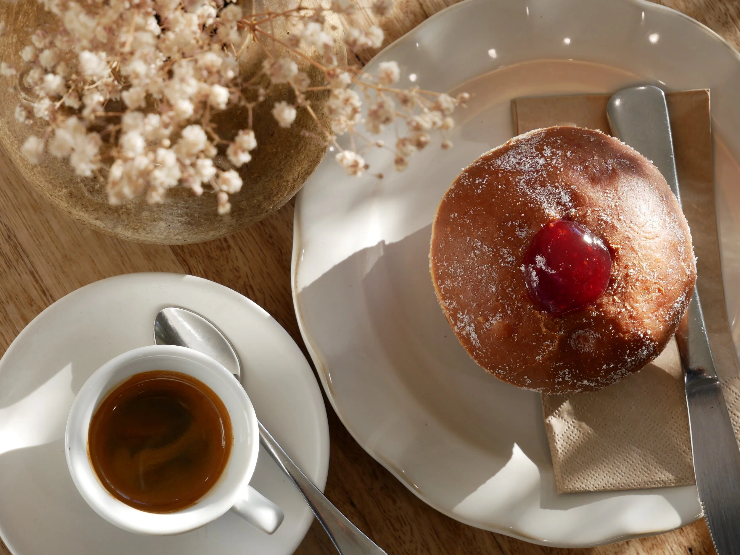 A white plate with a pastry topped with powdered sugar and a cherry, a cup of coffee, and a cup of tea, with a bouquet of white flowers on a wooden table.