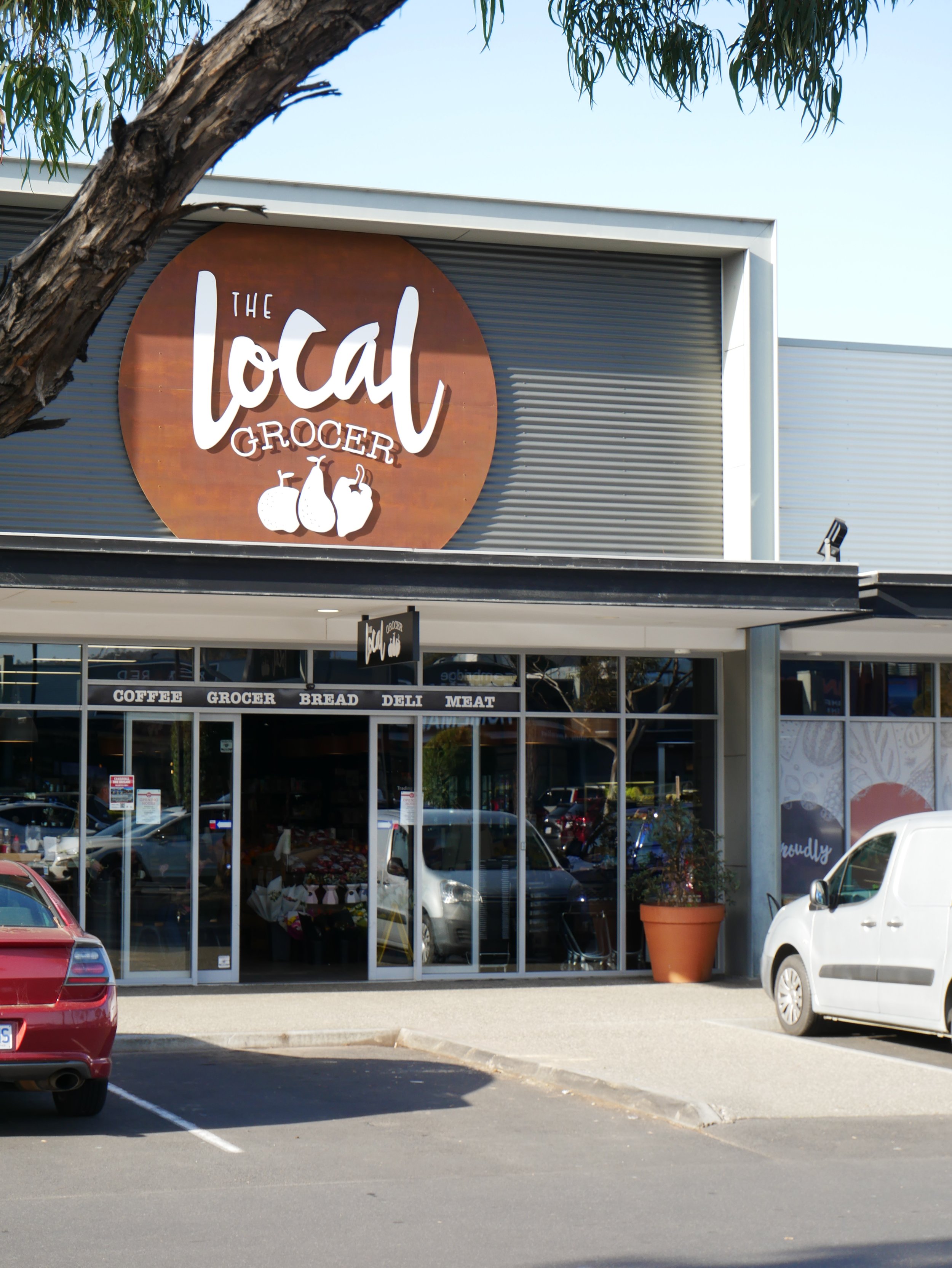 Front view of a grocery store with a sign reading 'The Local Grocer' and fruits on the sign. Glass doors and windows, parking lot with cars, and a large potted plant outside.
