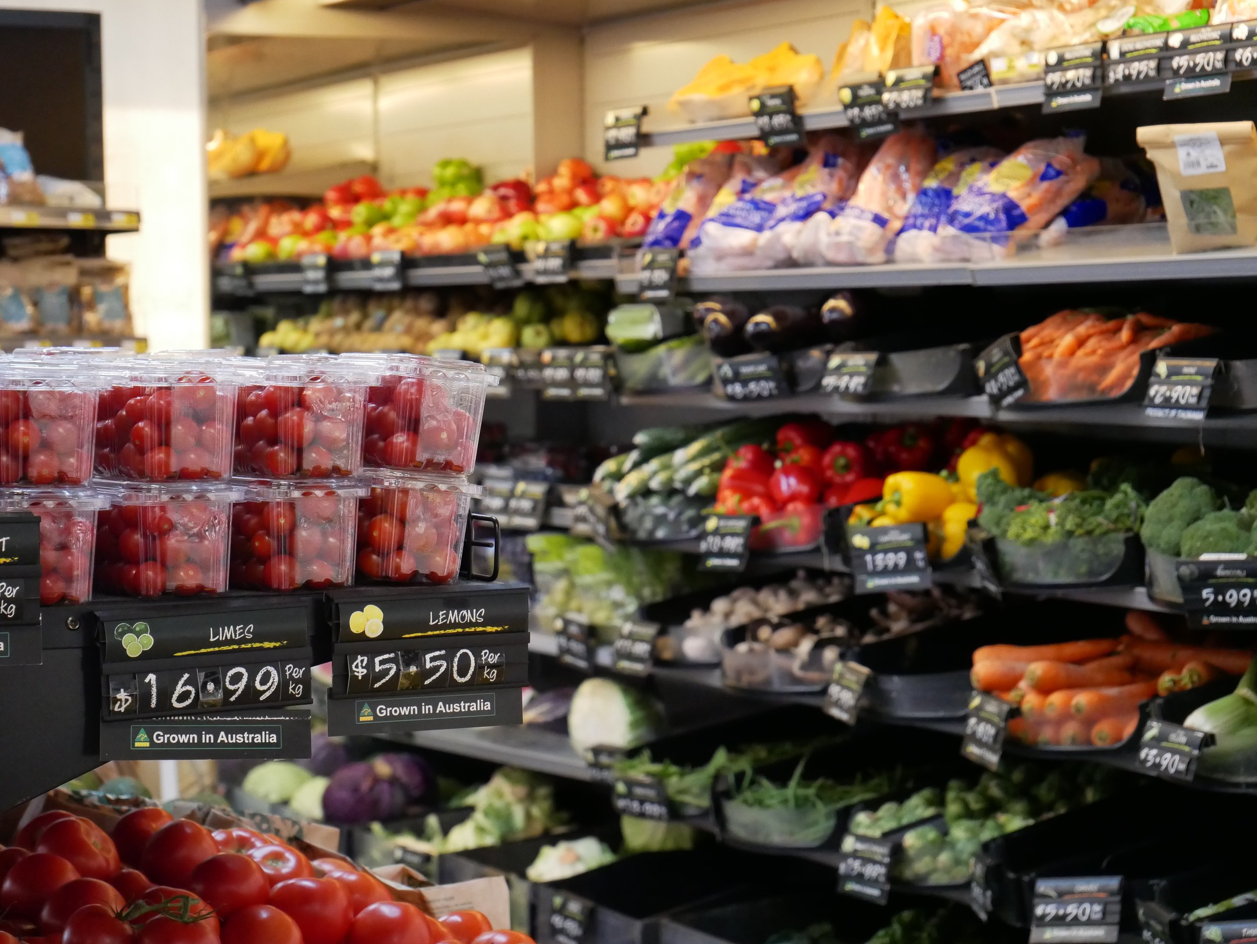 Grocery store produce section with fresh vegetables and fruits on shelves, including tomatoes in plastic containers and assorted vegetables like bell peppers, zucchini, and broccoli.