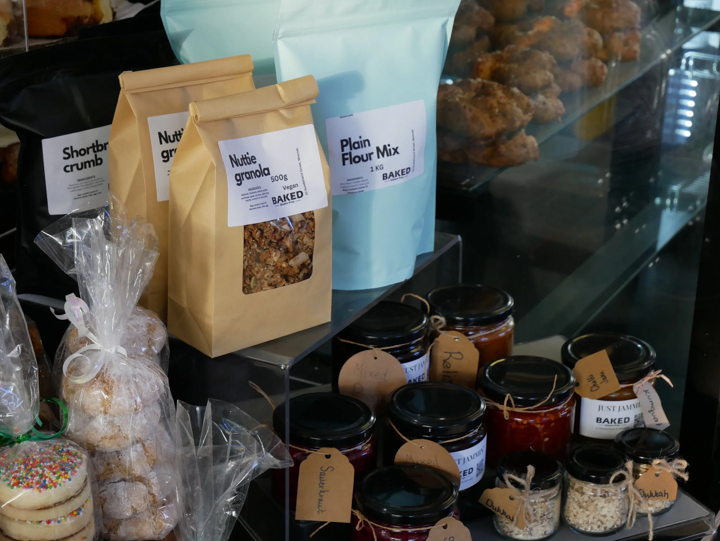 Assorted baked goods, including cookies in clear plastic bags, jars of jam, and packages of flour and granola, displayed on shelves.