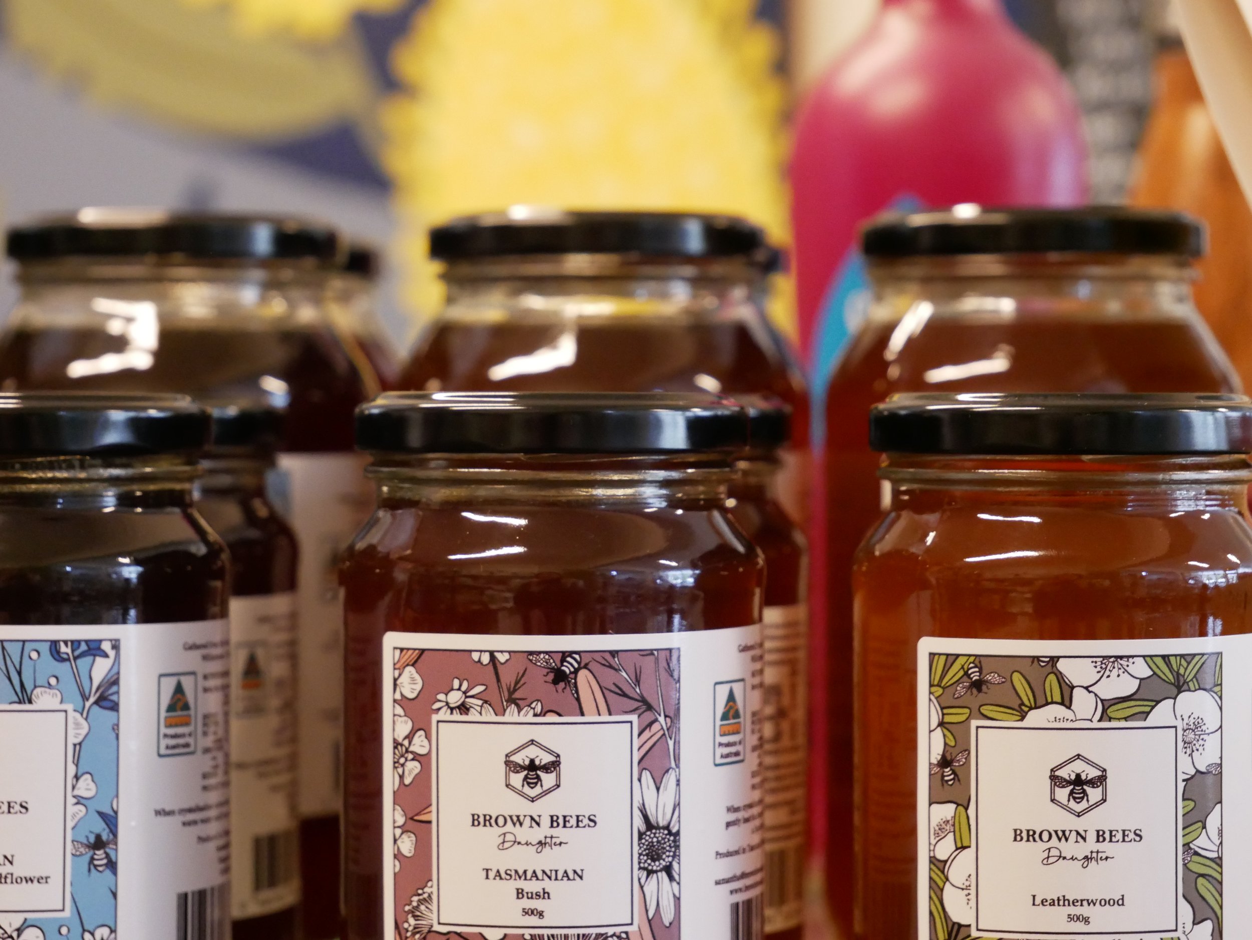 Close-up of six jars of honey with decorative labels depicting bees and floral designs, labeled as "Brown Bees", "Tasmanian Bush", and "Leatherwood", indicating different types of honey. In the background, there are blurred colorful objects.