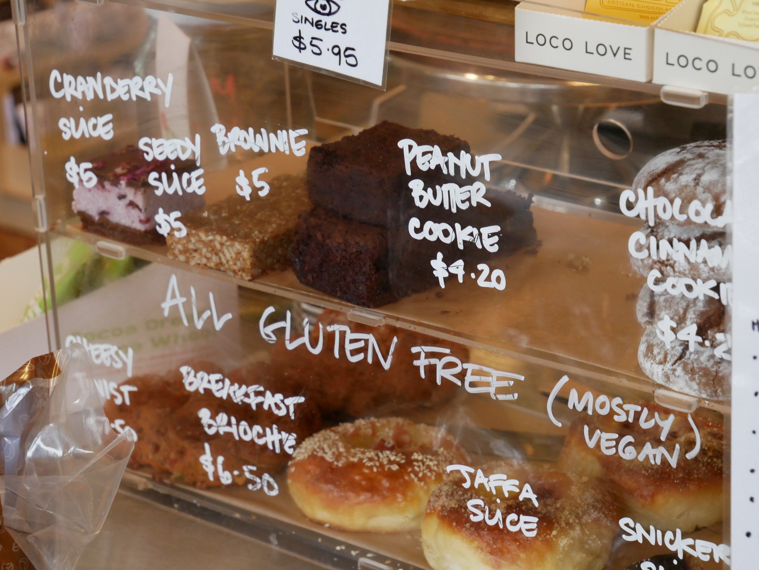 A display case with various baked goods, including slices of cake, with handwritten labels showing prices and flavors, such as cranberry, peanut butter, and chocolate cinnamon.