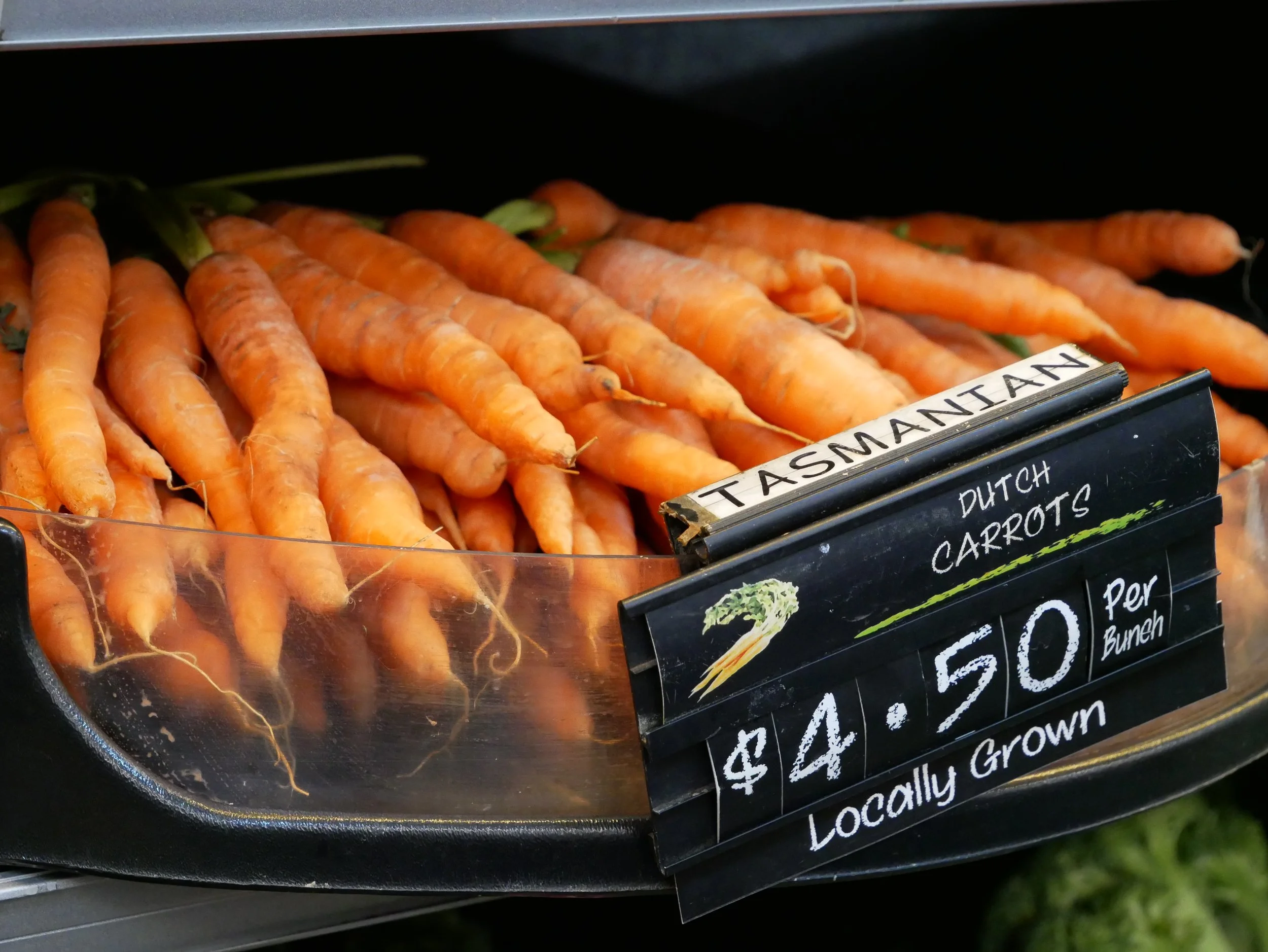 Fresh orange carrots with green tops on display at a grocery store, labeled as locally grown.