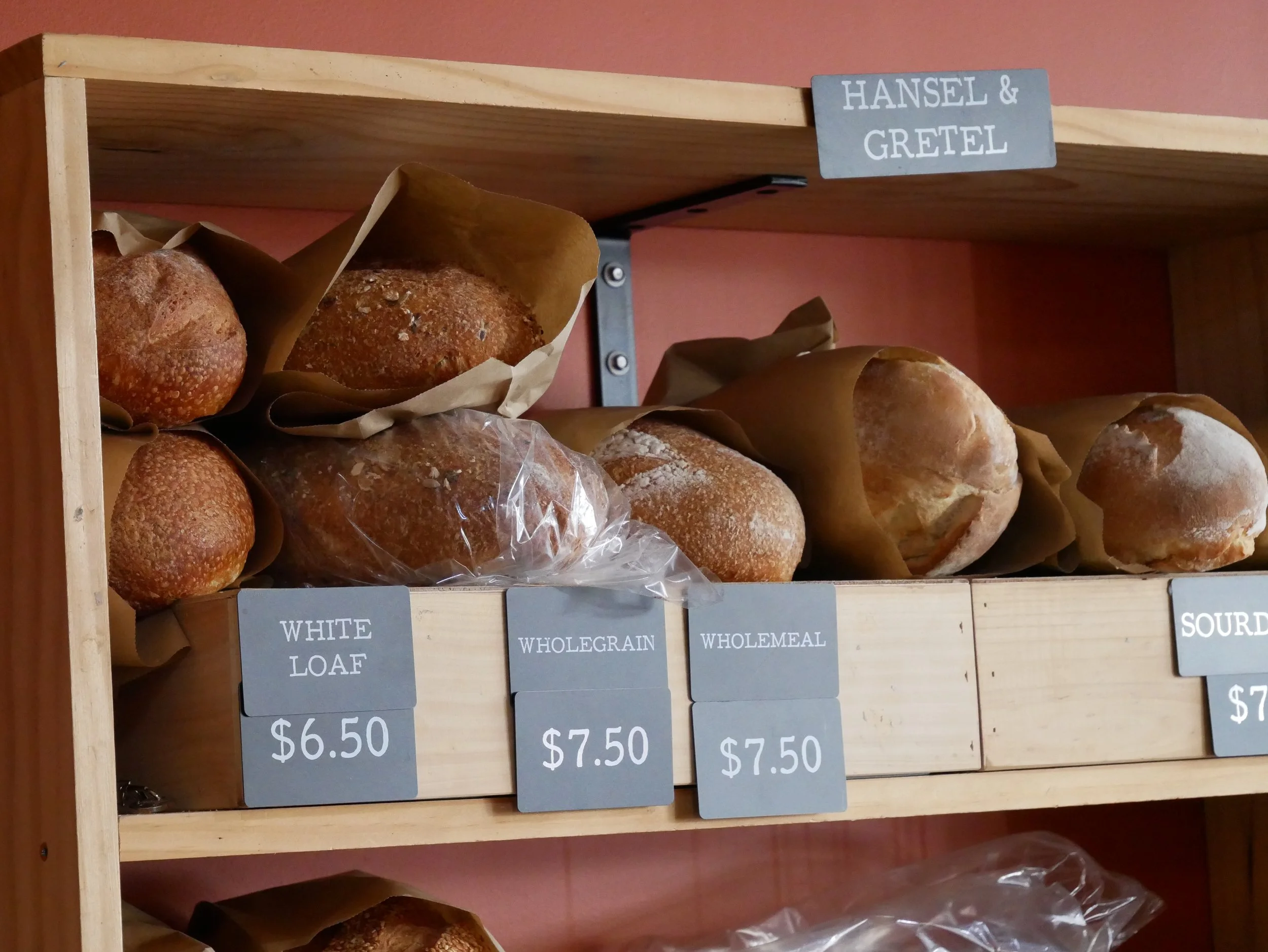 Shelves with various types of bread wrapped in paper and plastic, labeled as white loaf, wholegrain, wholemeal, and sourdough, with prices listed on a blackboard.