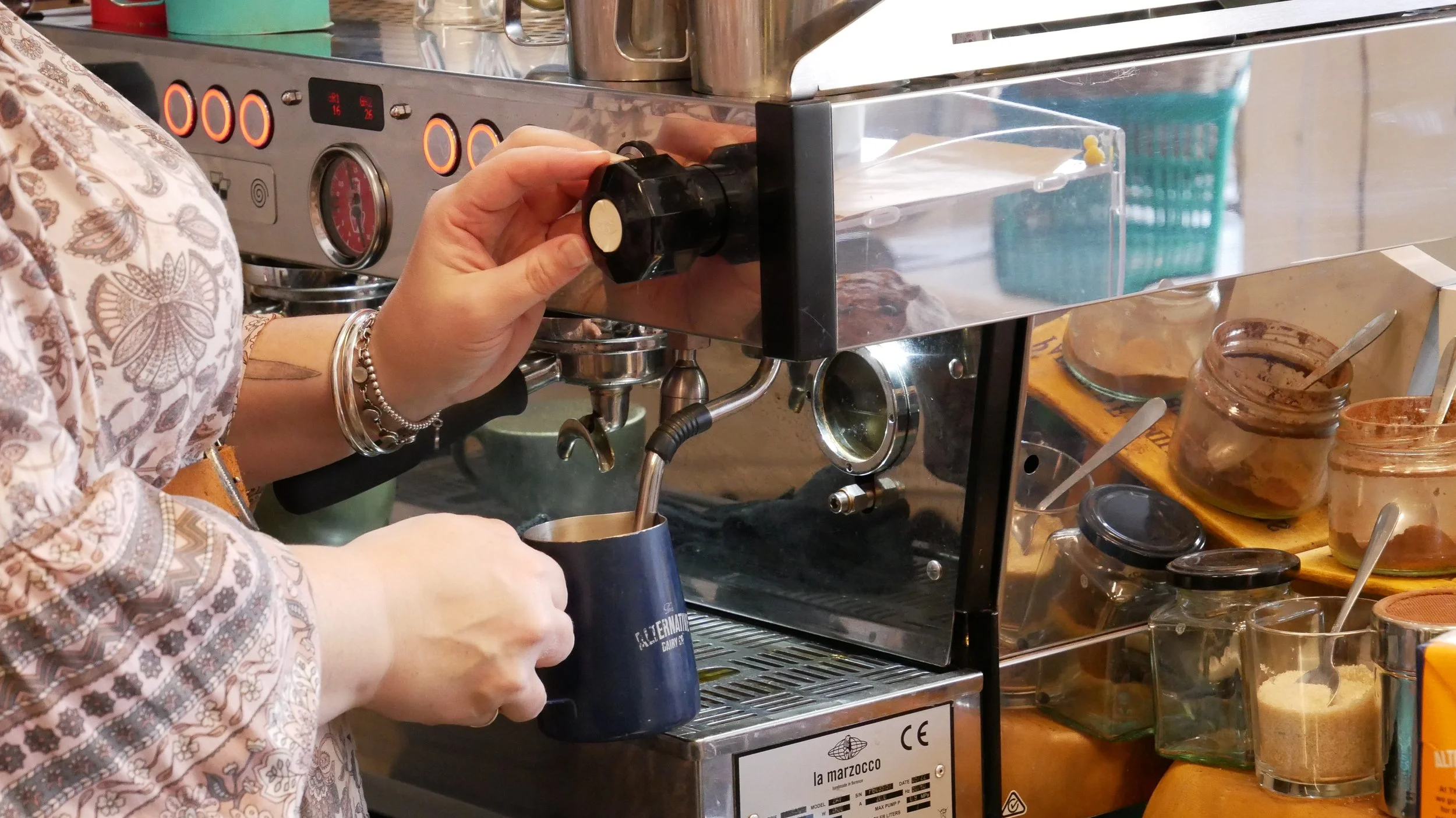 Person operating an espresso machine in a cafe, wearing a patterned sleeve, pouring hot water or steam into a cup.