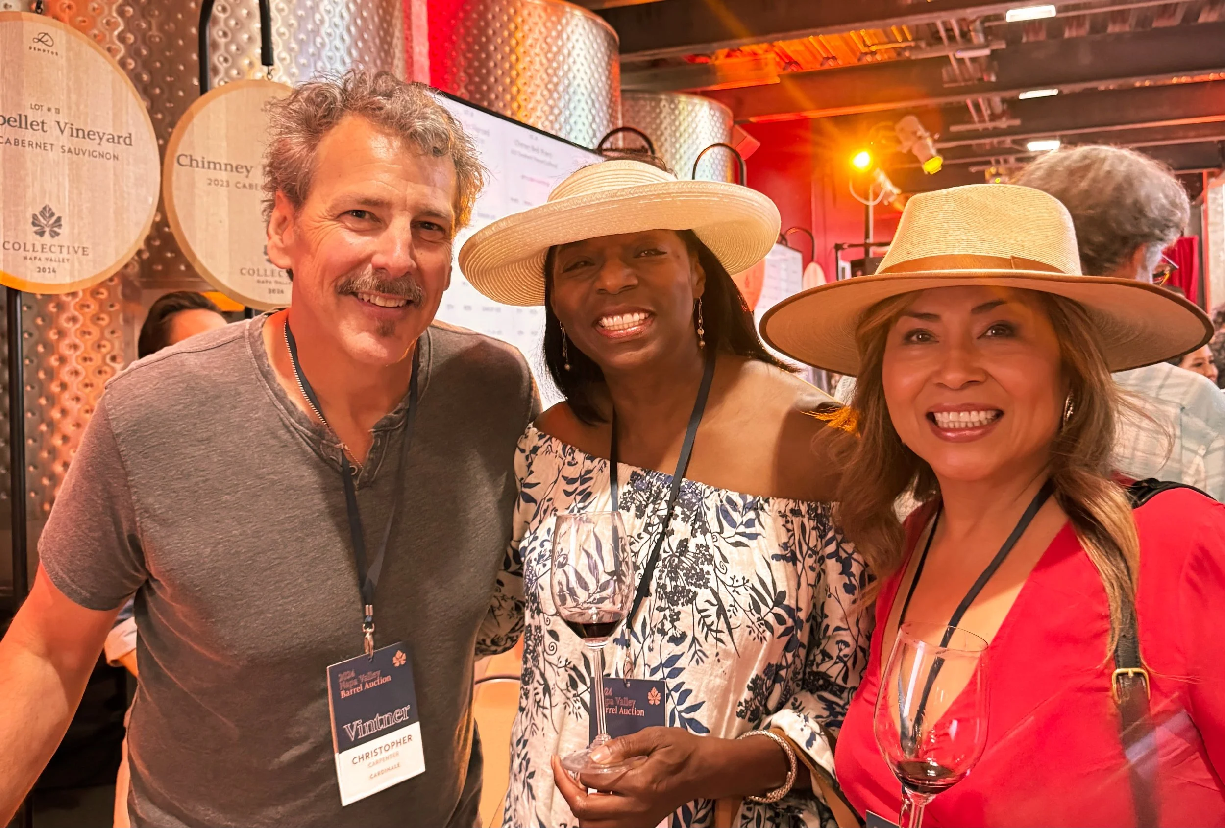 Two people in sun hats holding glasses of red wine in a barrel room hung with vineyard lot signs