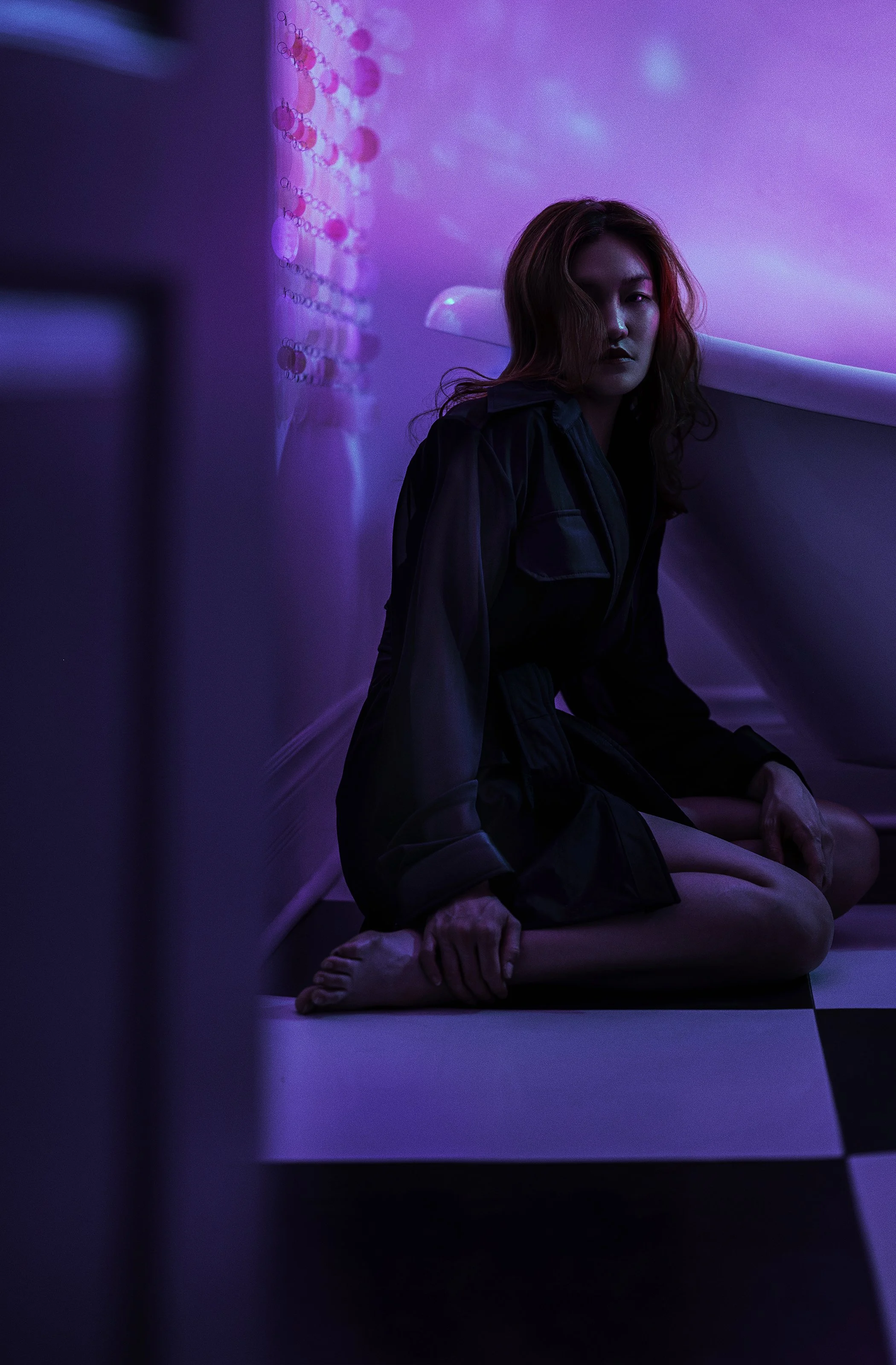 A woman with red hair wearing a dark jacket sitting on a black and white checkered floor in a dimly lit room with colorful neon lights and abstract wall art.