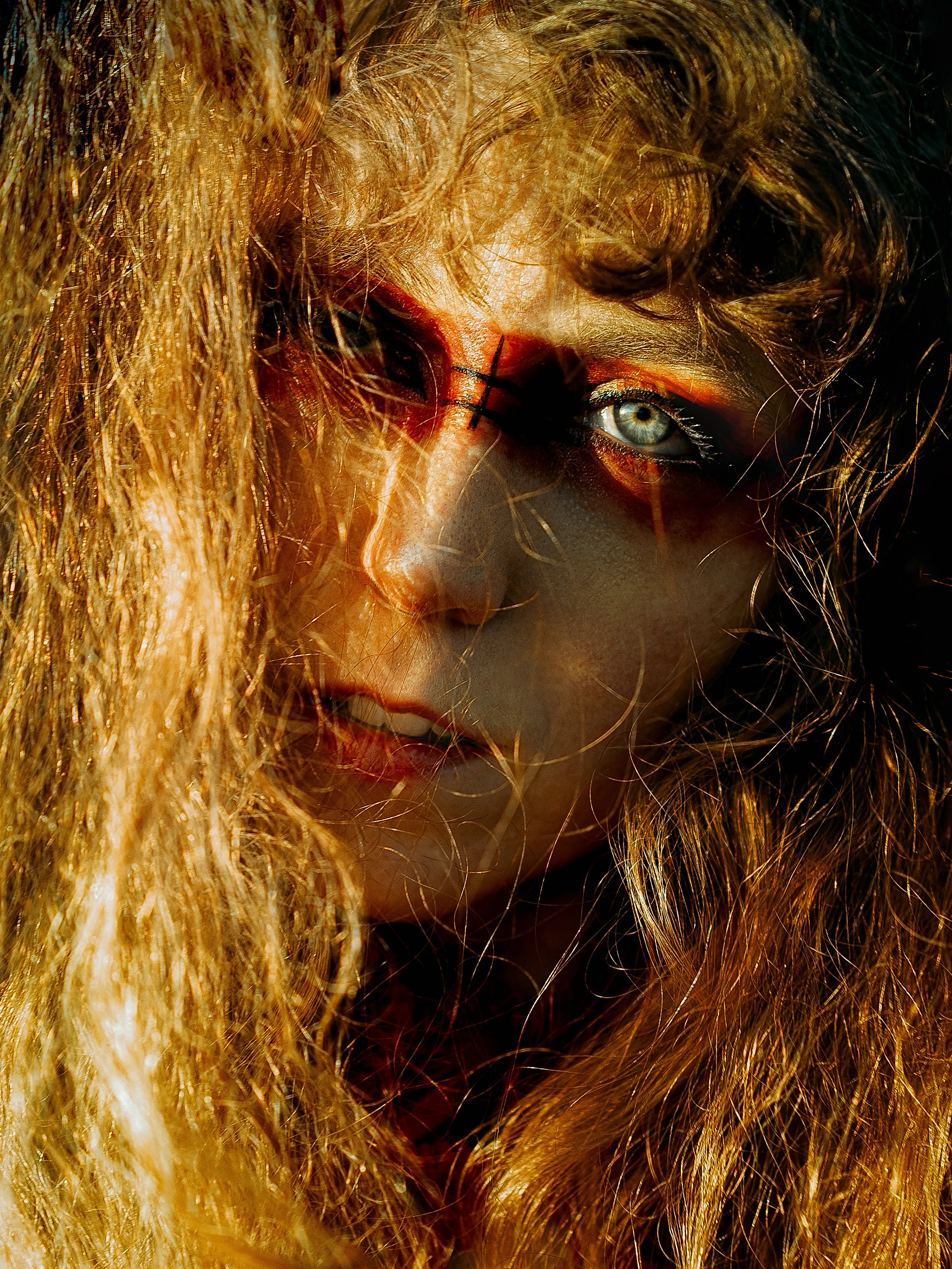 Close-up of a woman with curly blond hair, intense eye makeup, and striking blue eyes, illuminated by dramatic lighting.