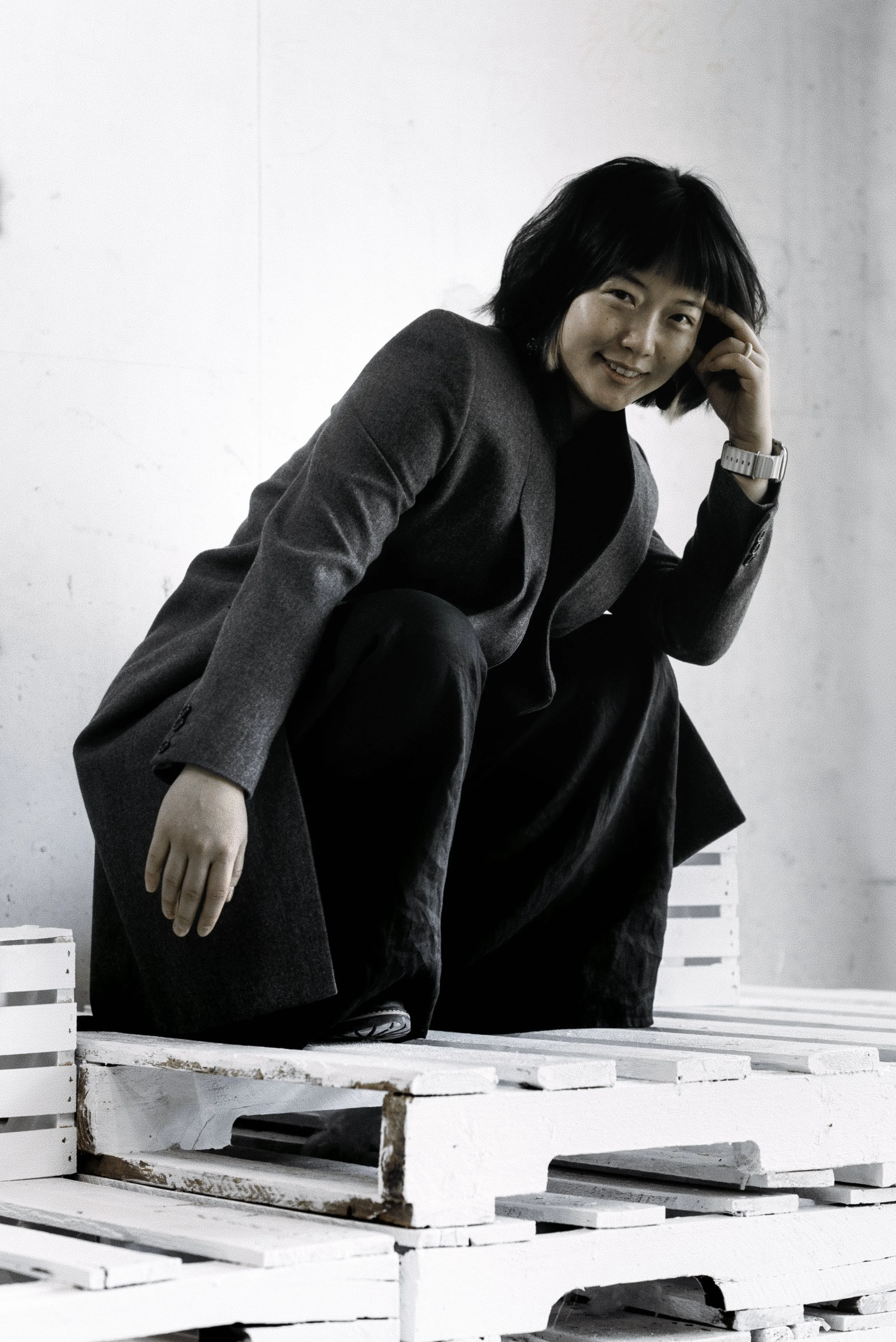 A woman squatting on white wooden pallets in a minimalistic, industrial-style setting, wearing a gray blazer and black trousers, smiling at the camera.