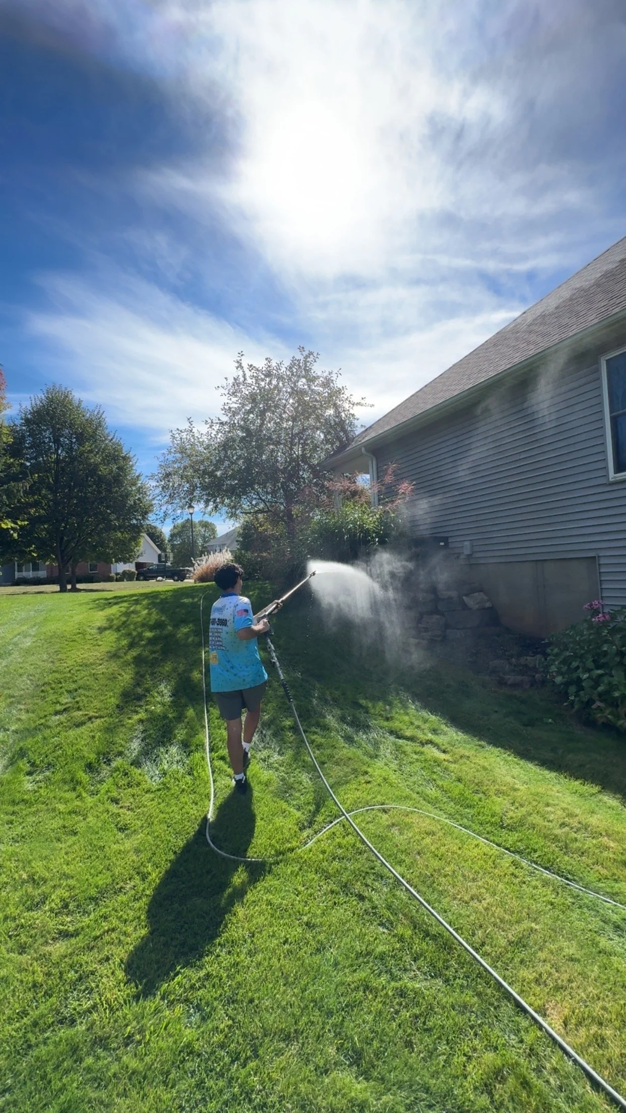 A boy wearing a blue sports jersey and gray shorts is using a garden hose for pressure washing the side of a house on a sunny day in a green, suburban backyard.