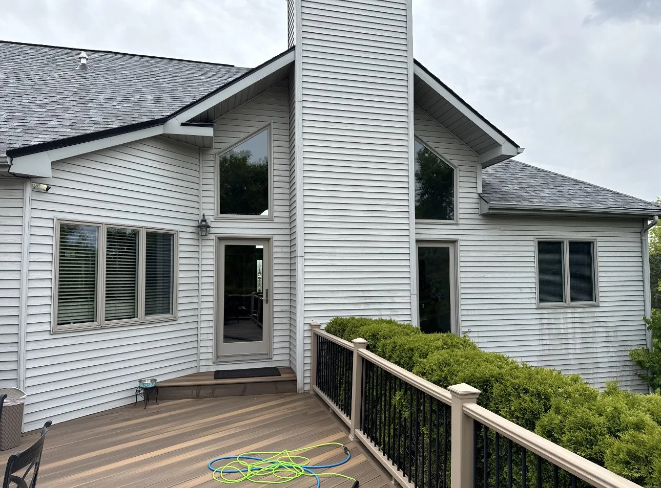 Back view of a multi-story house with white siding, large windows, and a wooden deck with a black railing. There is a small green shrub and garden hose on the deck.