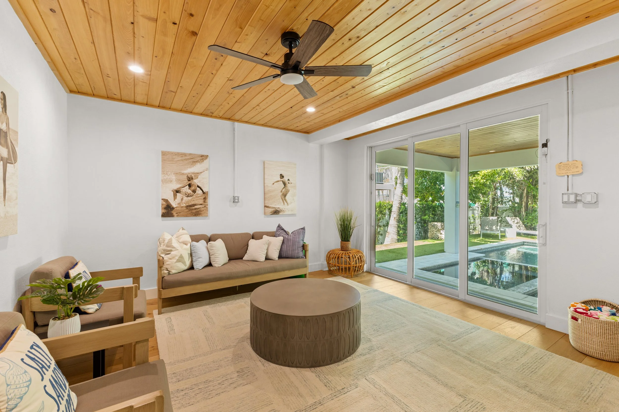 Photography of a living room with a wooden ceiling, ceiling fan, white walls decorated with beach-themed artwork, large sliding glass doors leading to an outdoor pool area.
