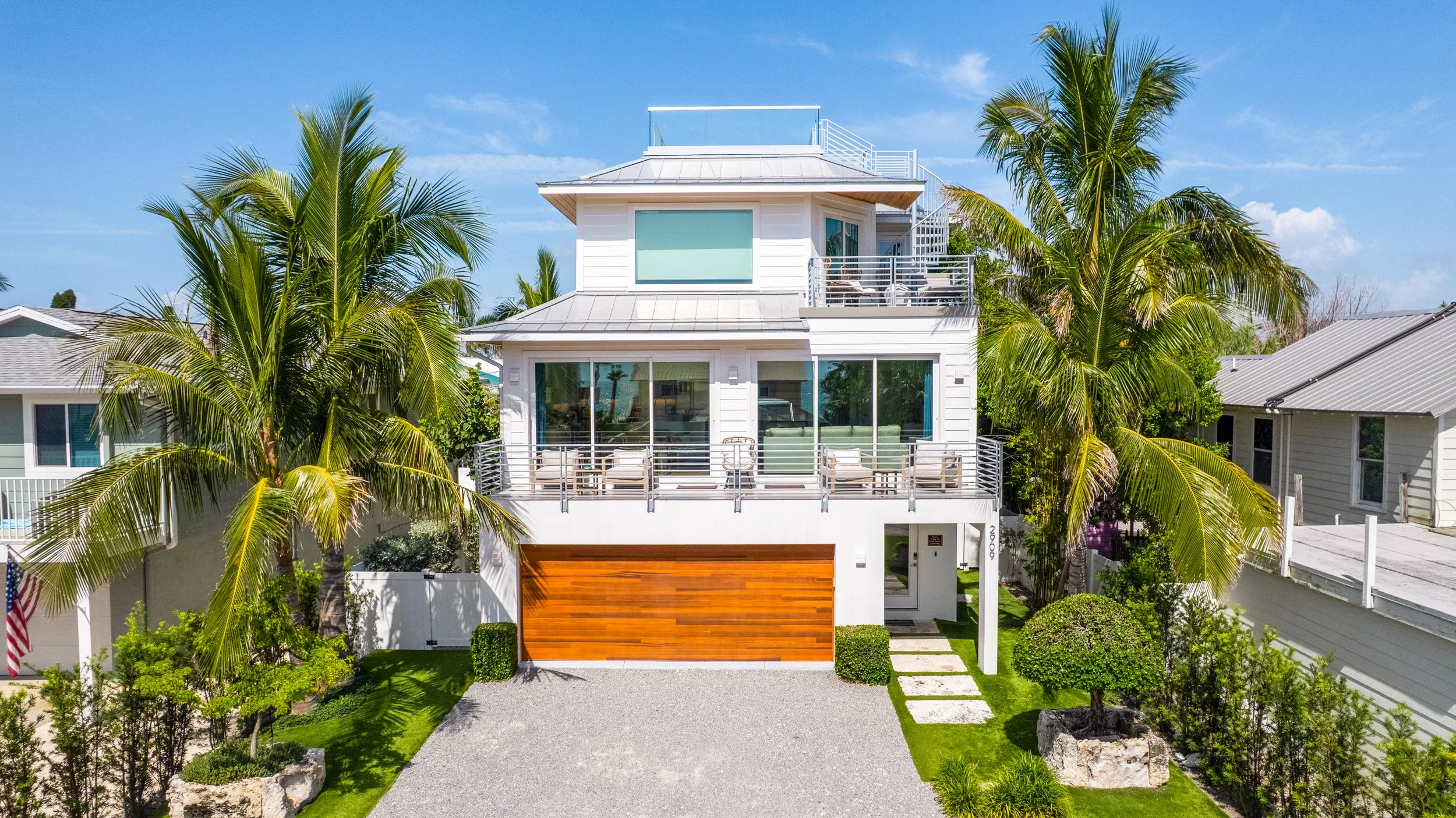 A modern multi-story house with a white exterior, large windows, and balconies, surrounded by palm trees and a landscaped yard with rocks, bushes, and a gravel pathway located in Bradenton, Florida