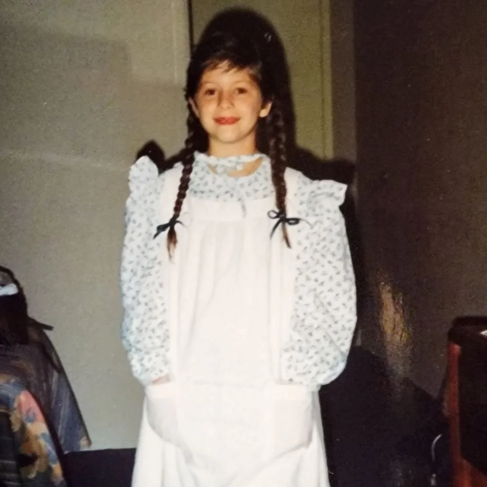 A white 10 year old girl with brown hair in braids is wearing a 19th century dress with a pinafore in a hotel room.