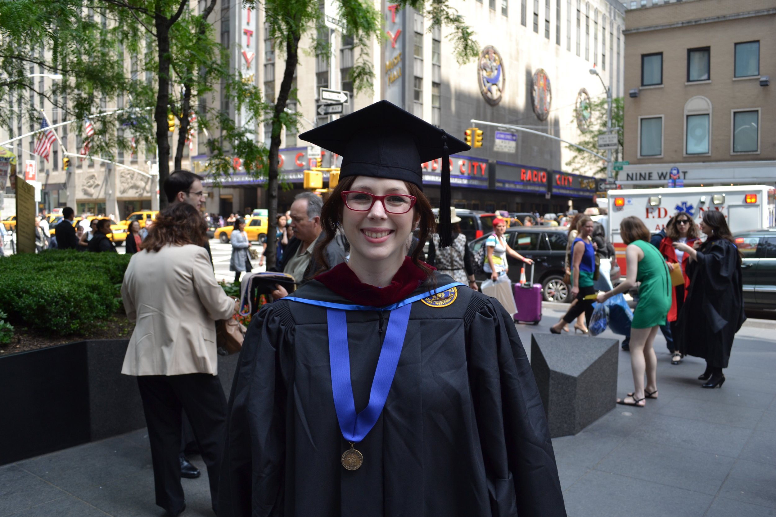 A woman in a graduation cap and gown stands in front of Radio City Music Hall