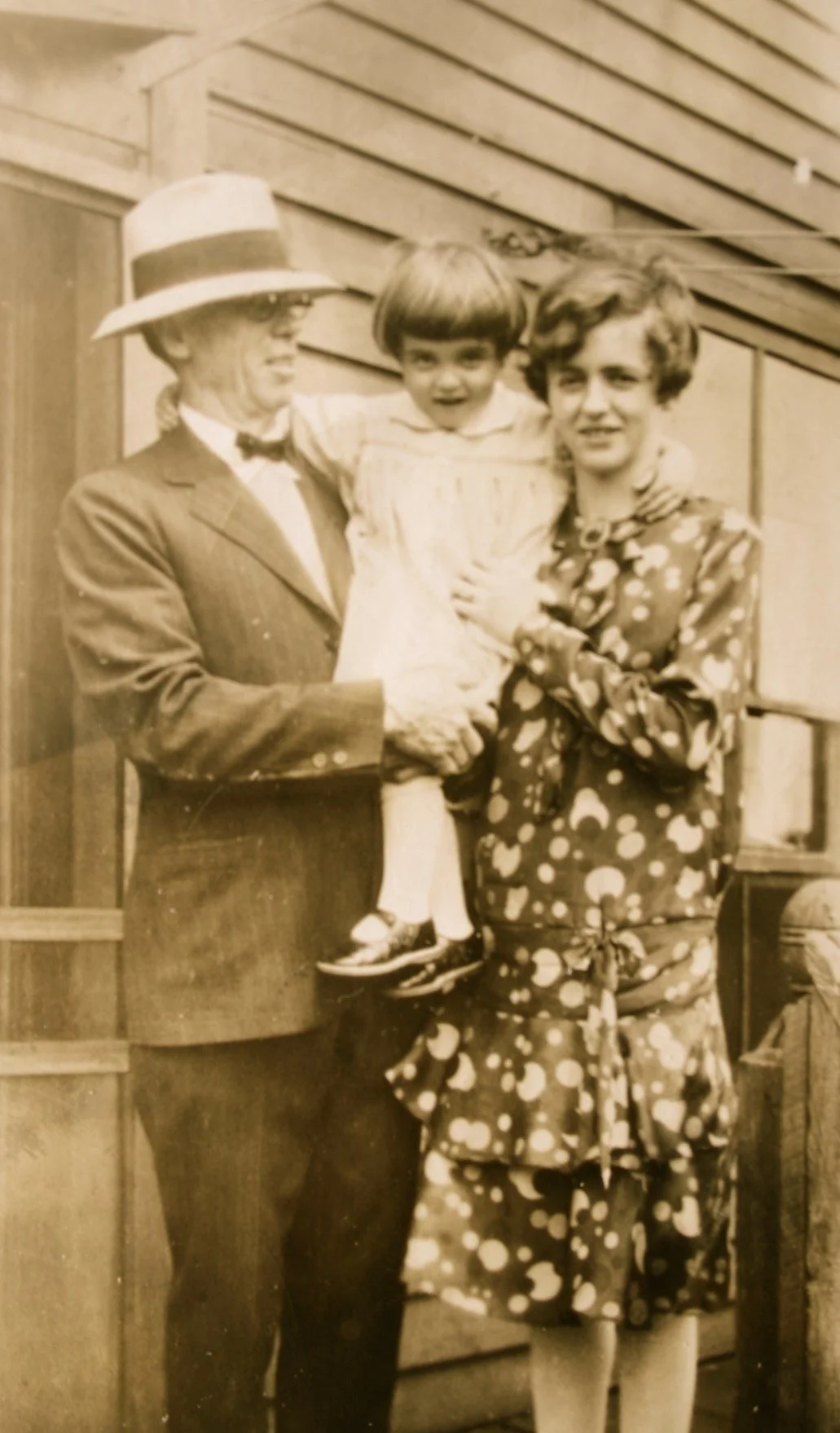 An older man in a straw hat and suit holds a toddler with a young woman in a dress standing next to them in an historical photo from around 1930