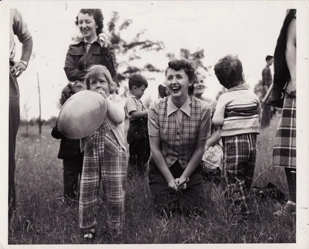 A black and white photo of a woman kneeling outdoors in a vest and shortsleeved button up, laughing, while a child holds a balloon and others gather nearby. Approximately 1940s