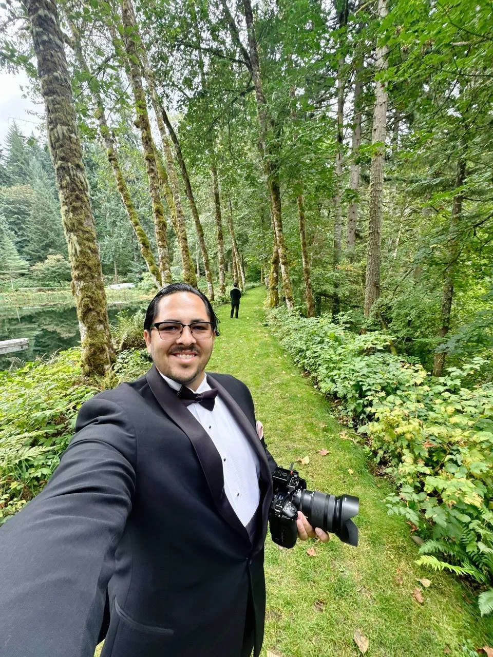 Male Videographer in a tuxedo standing in a forest with a groom waiting for the first look.