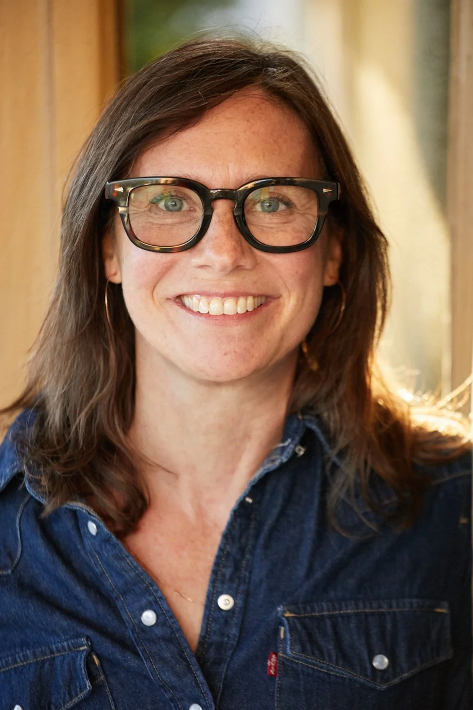 A woman with brown hair and glasses smiling at the camera, wearing a denim shirt.