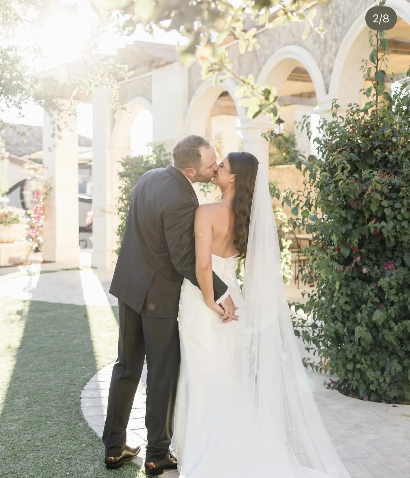 A wedding couple sharing a kiss outdoors, with sunlight filtering through trees, archways, and lush greenery around them.