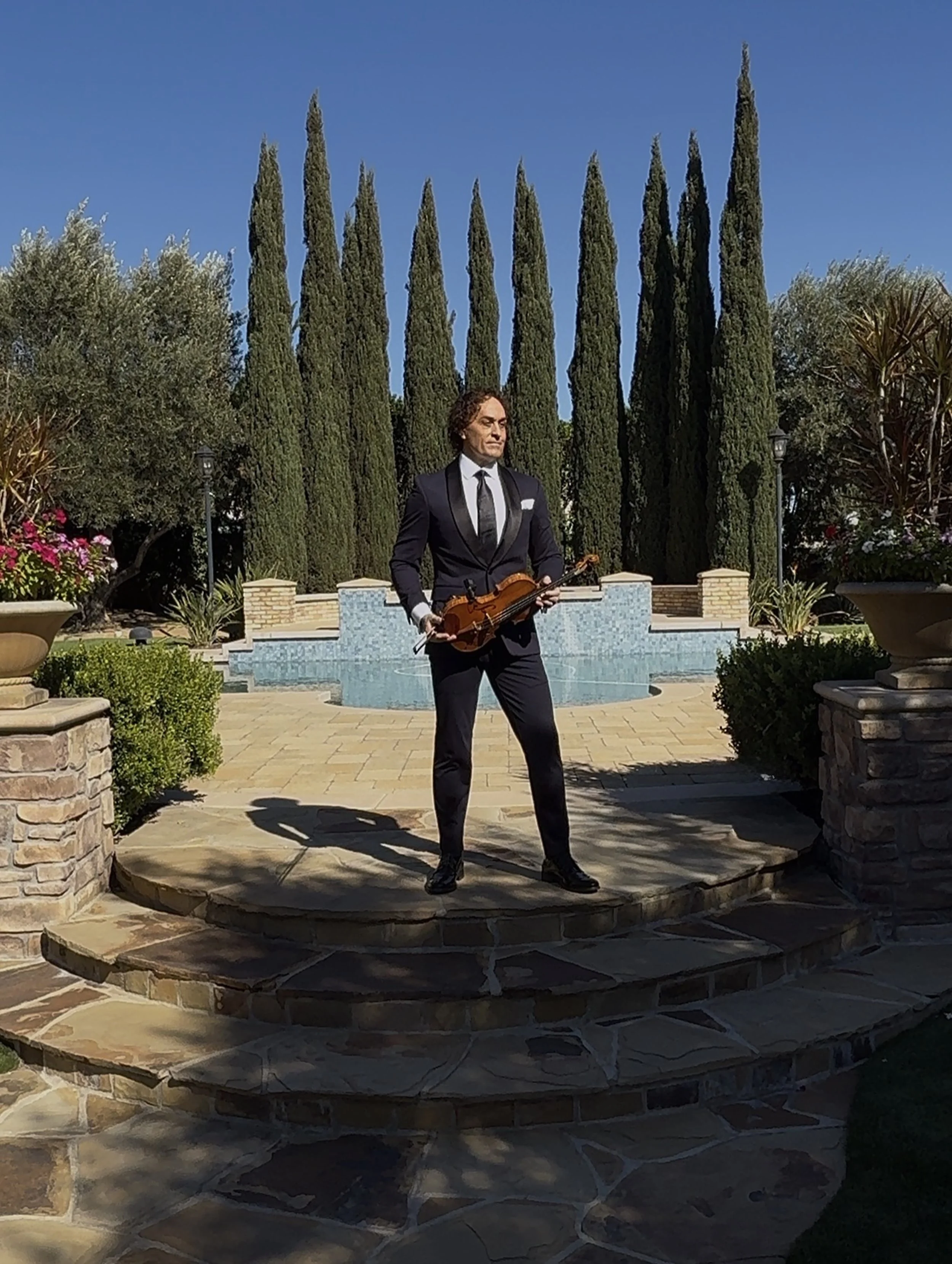 Tim Espinosa in a black tuxedo holding a violin and bow, standing on stone steps outdoors with tall cypress trees and a water fountain in the background.