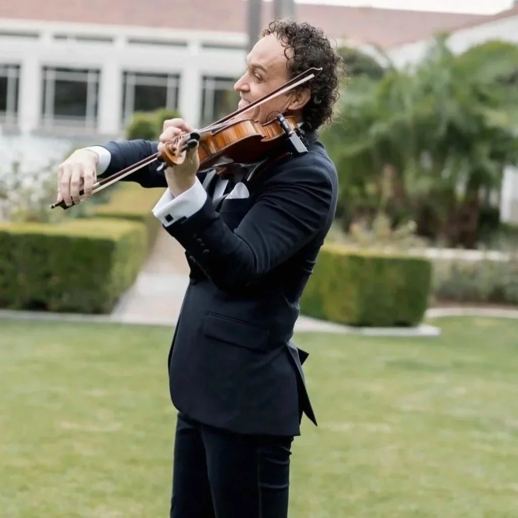 A man in a black tuxedo playing a violin outdoors on a lawn with a modern building and green trees in the background.
