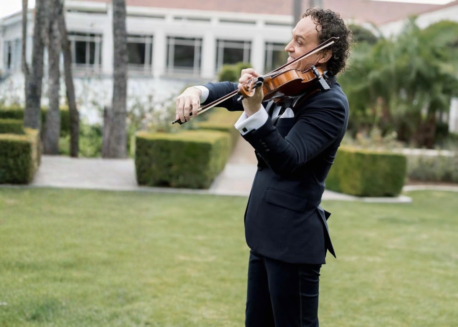 Tim Espinosa performing violin on a manicured estate lawn lined with hedges and palms
