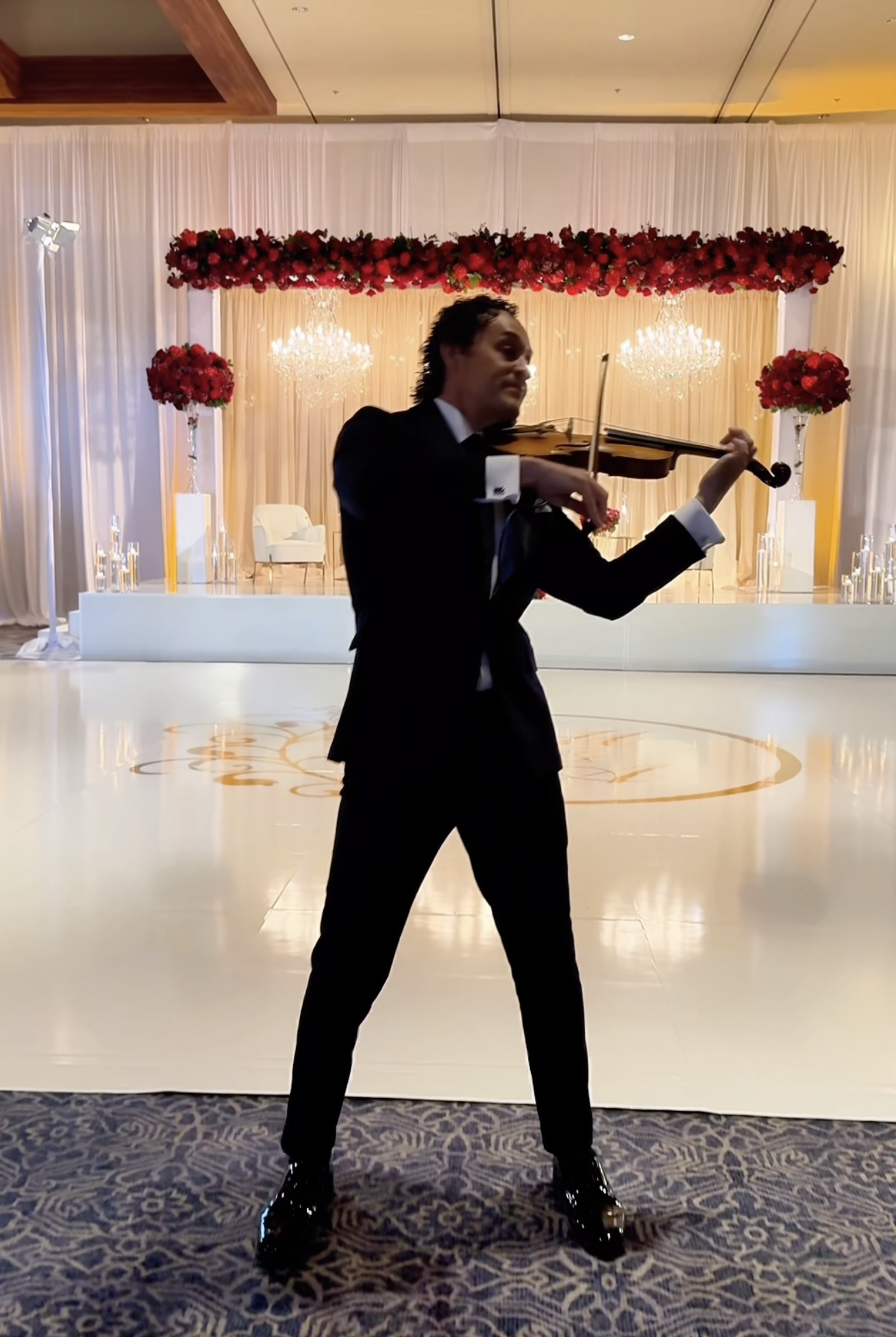 Tim Espinosa in a black tuxedo playing a violin at a formal event with a decorated stage featuring red flowers, chandeliers, and white drapery in the background.