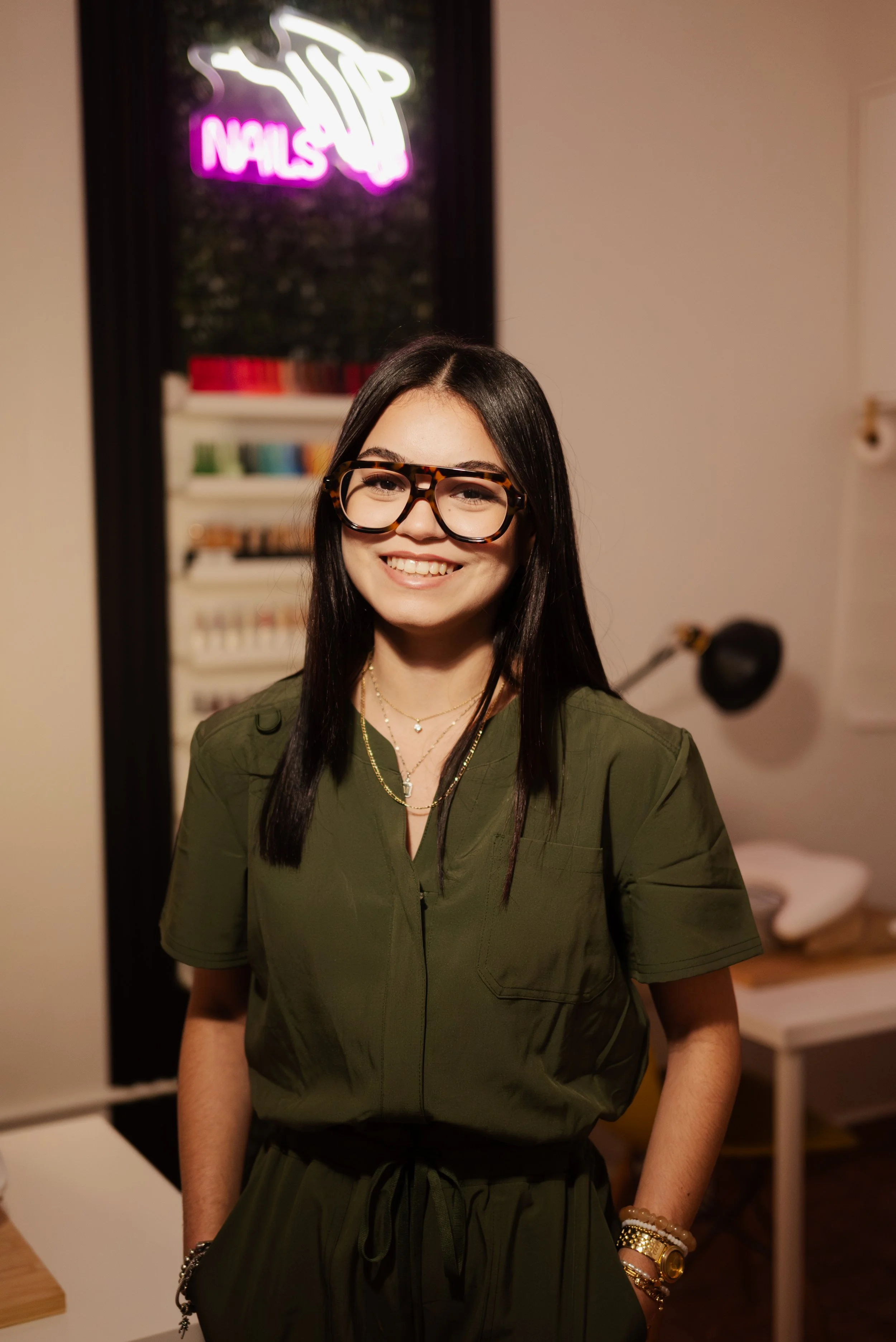 A young woman with long black hair and glasses smiling at the camera in a nail salon, with a neon sign that says 'Nails' in the background.