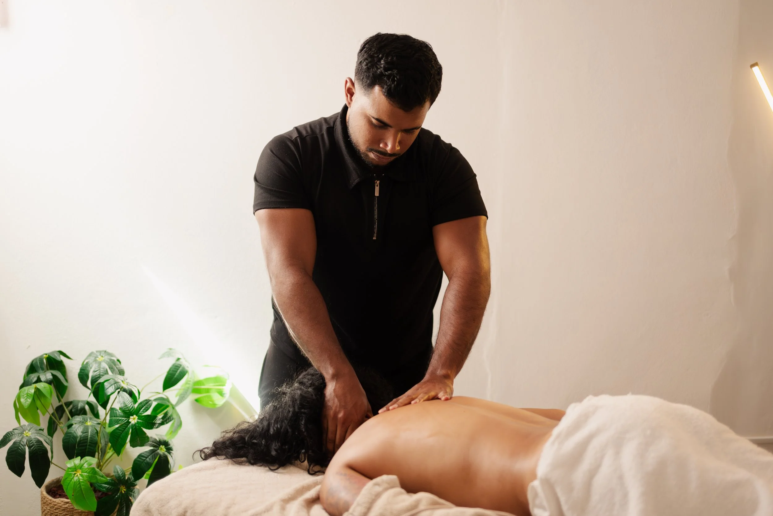 A man giving a massage to a woman lying face down on a massage table in a room with a white wall and a green plant.