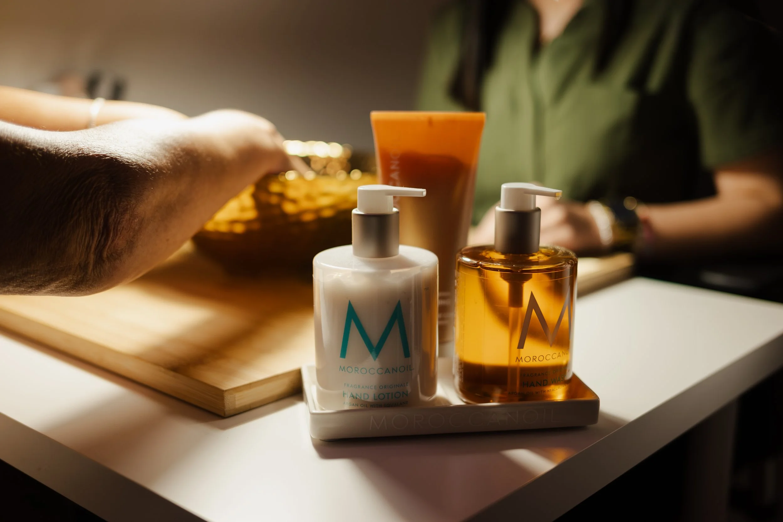 Two bottles of Moroccanoil hand lotion and hand wash on a white tray, with a person in a green shirt in the background, on a white table with a wooden tray.