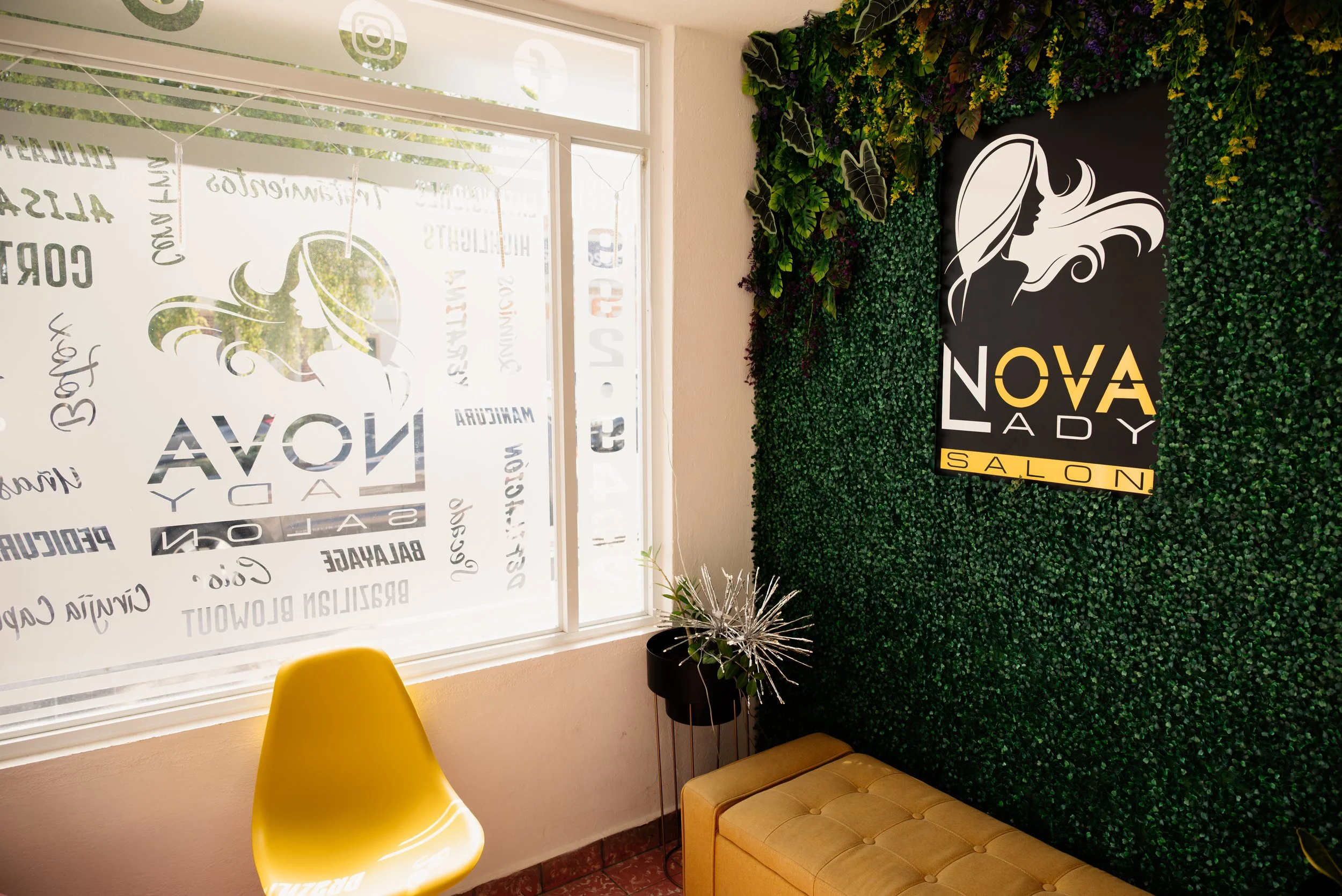 Interior of Nova Lady Salon with a large green leafy wall, a black and yellow sign, a yellow chair, and a small side table with plants. The window has the salon's name and services listed.
