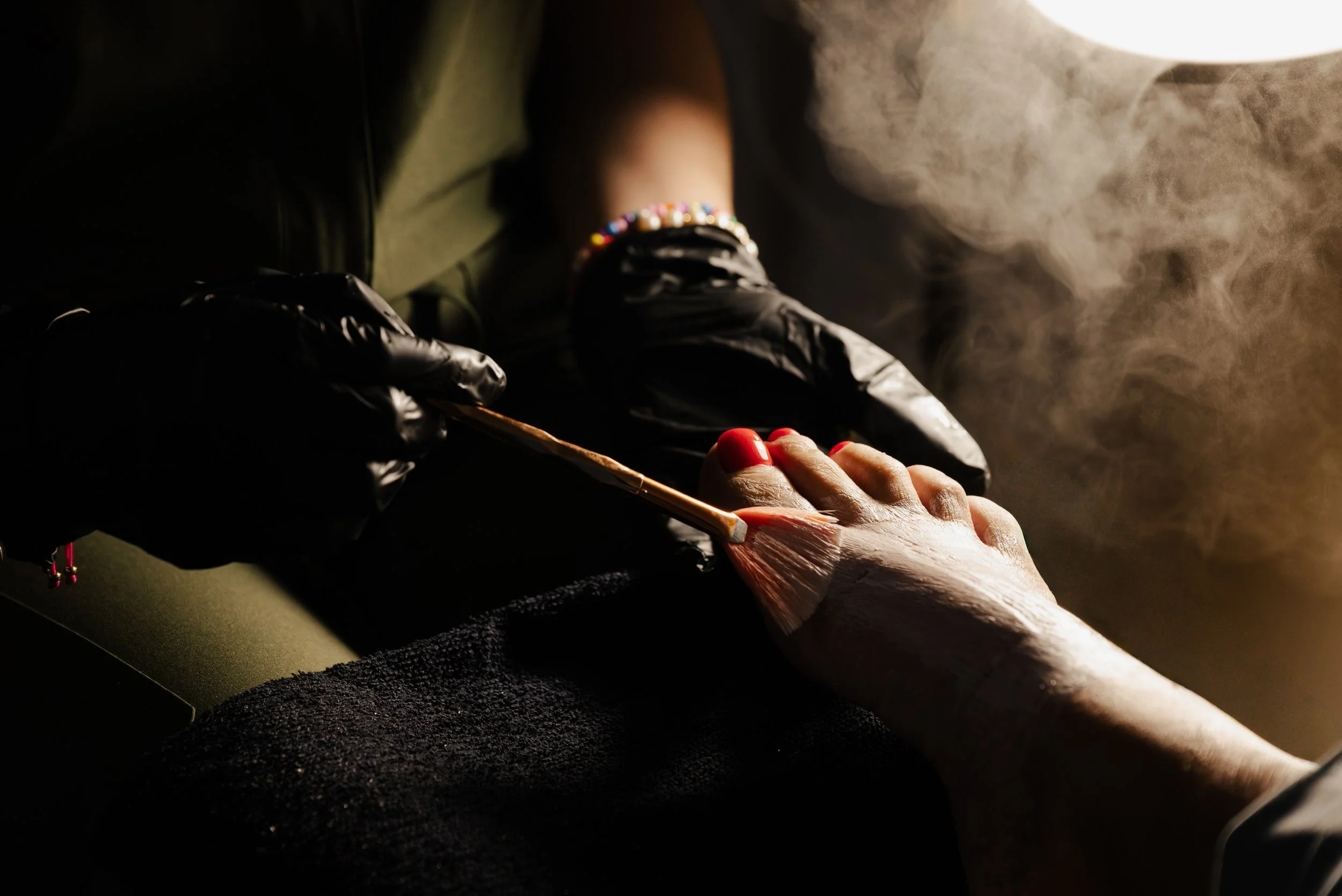 A tattoo artist in black gloves is applying a tattoo to a person's foot with a brush, with visible steam or smoke in the background.