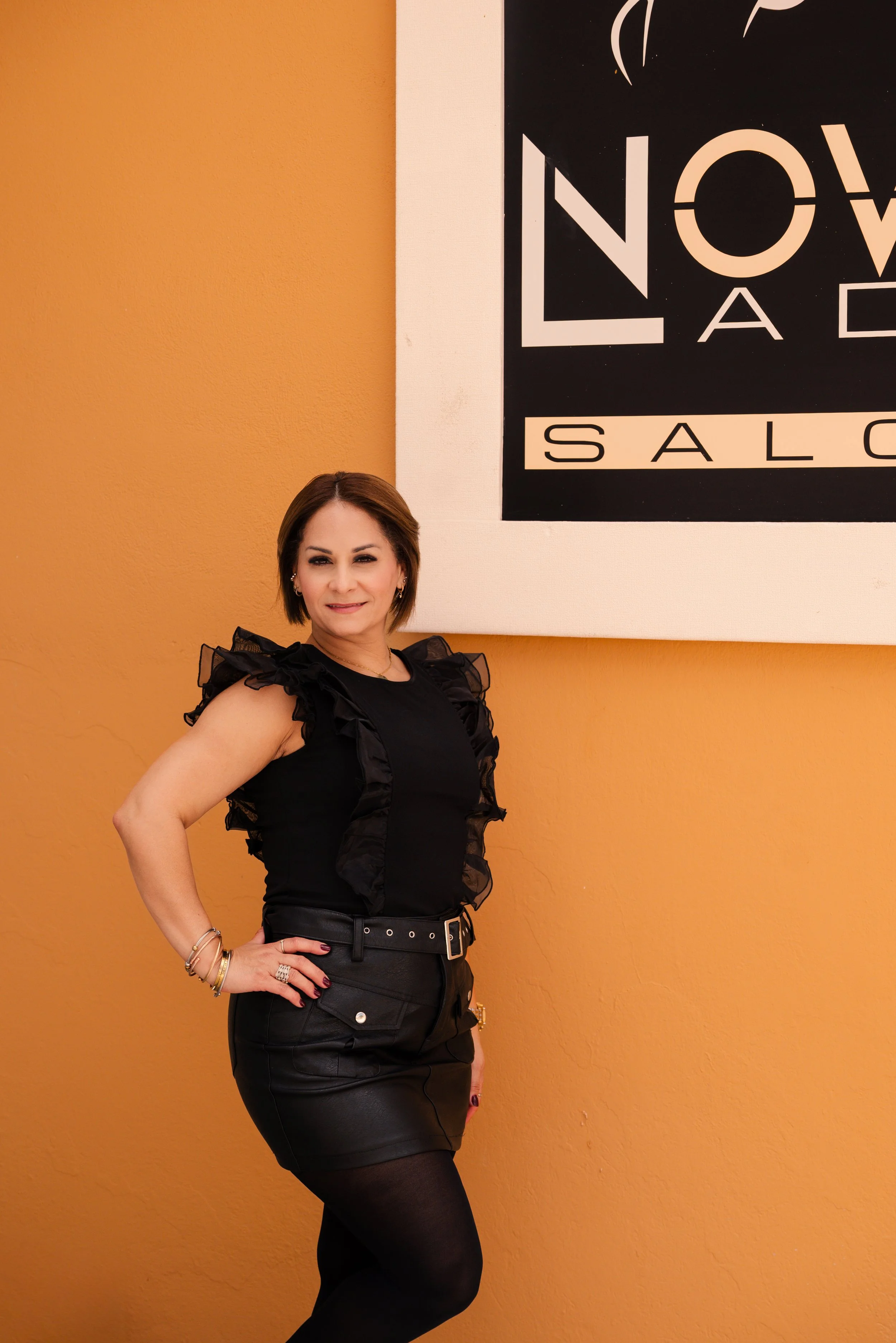 A woman with short brown hair wearing a black sleeveless ruffled top, black leather skirt, and black tights standing in front of an orange wall with a large black and white sign.