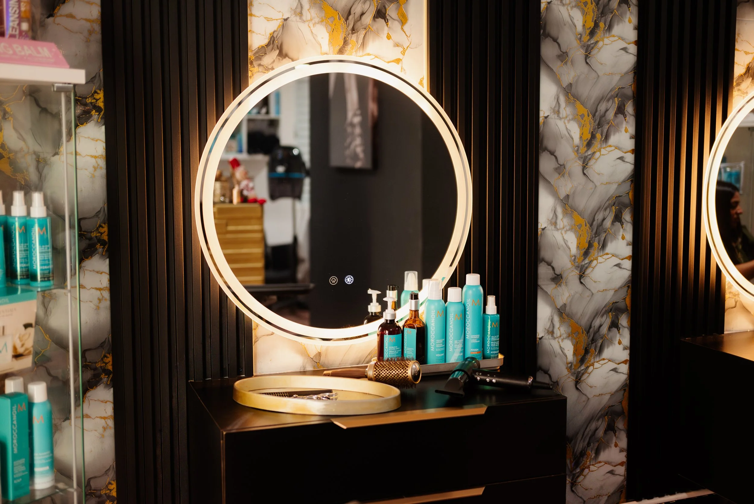 A modern vanity mirror with LED lighting on a marble wall backsplash, surrounded by haircare and skincare products on a black countertop in a salon or bathroom.