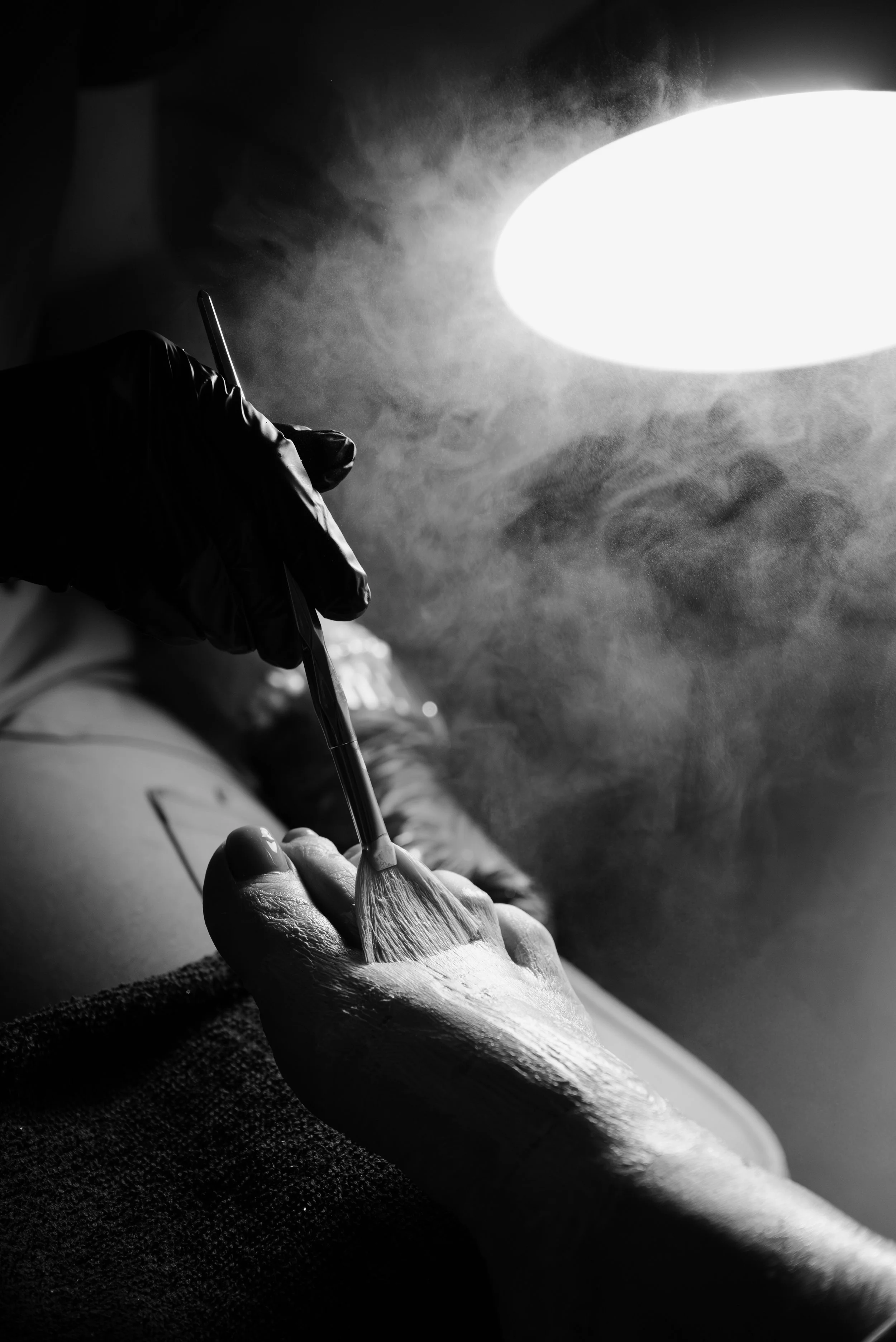 A person wearing black gloves is painting a woman's shoulder with a brush under a bright artificial light with smoke or mist around.