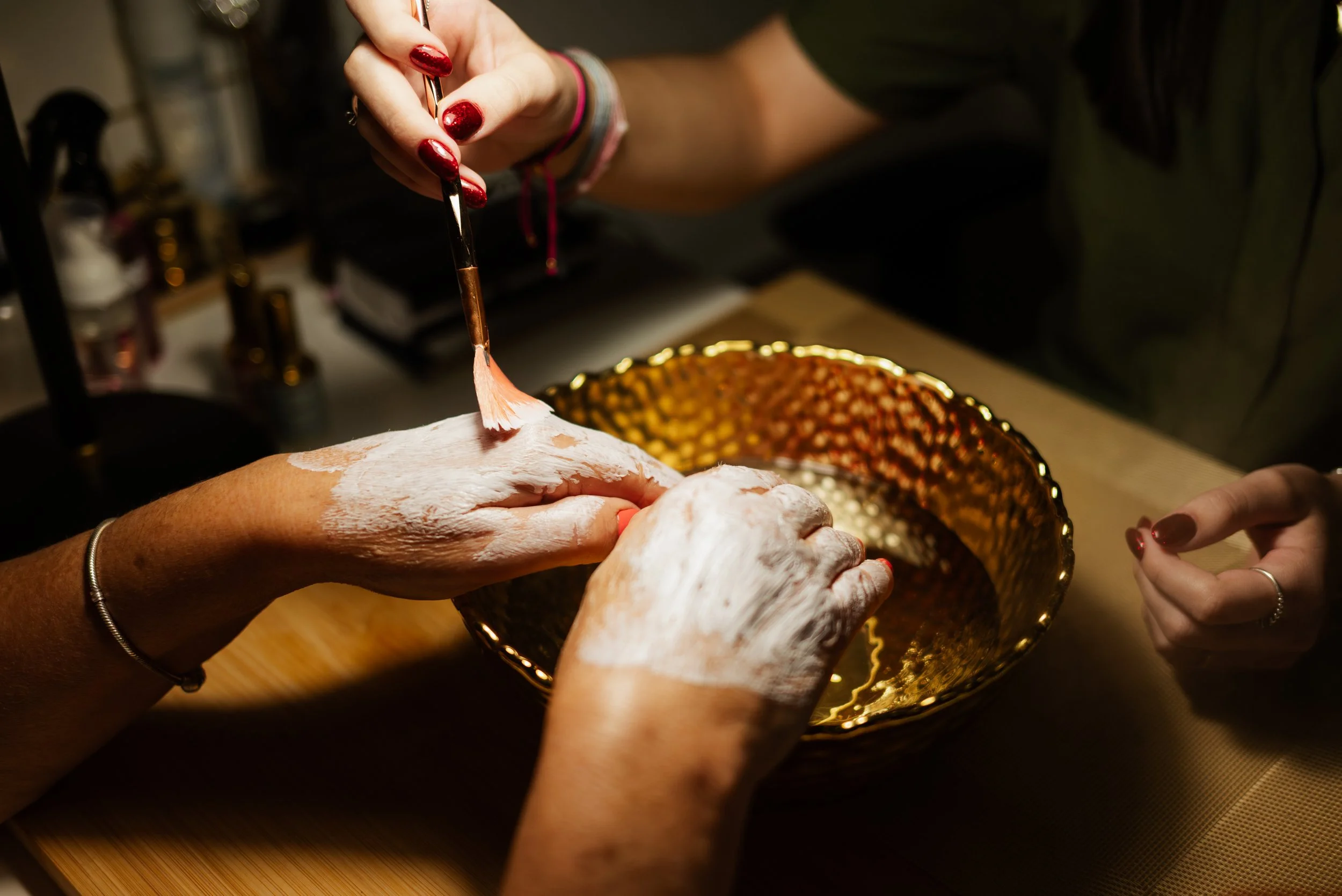 Person applying white face mask to another person's hand with a small brush, on a wooden table.