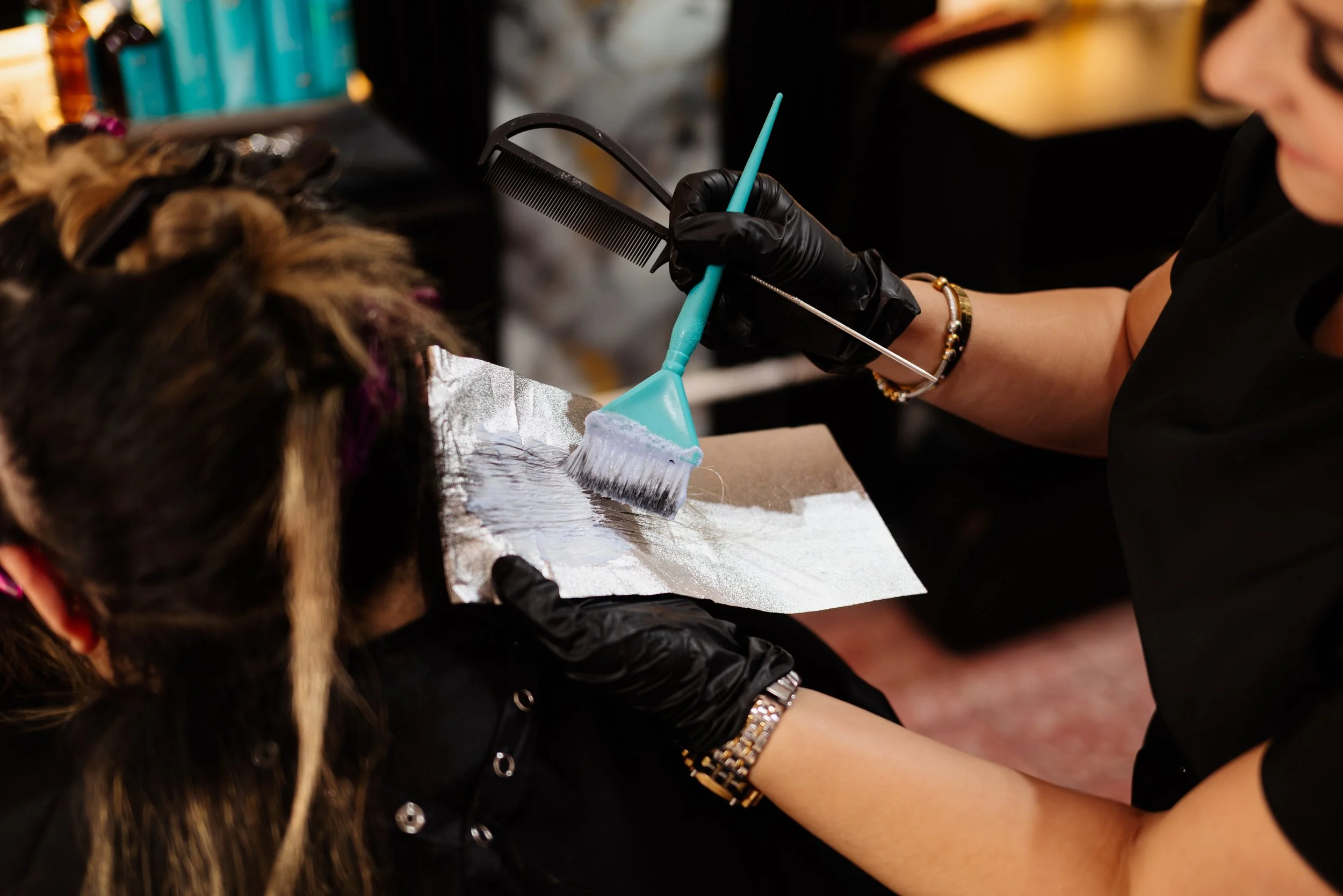 A hairstylist applying hair dye to a client's hair with a brush, wearing black gloves.