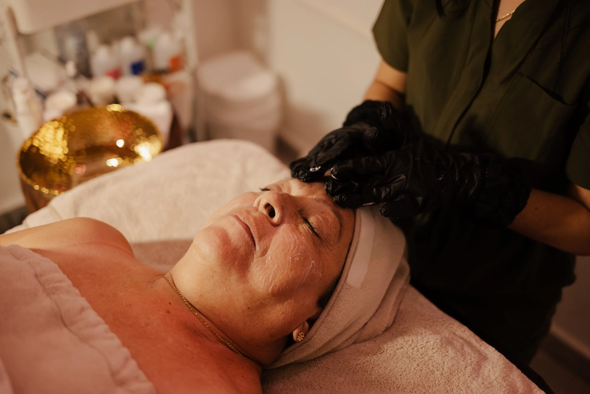 A woman with closed eyes receiving a facial treatment from an esthetician wearing black gloves in a spa room.