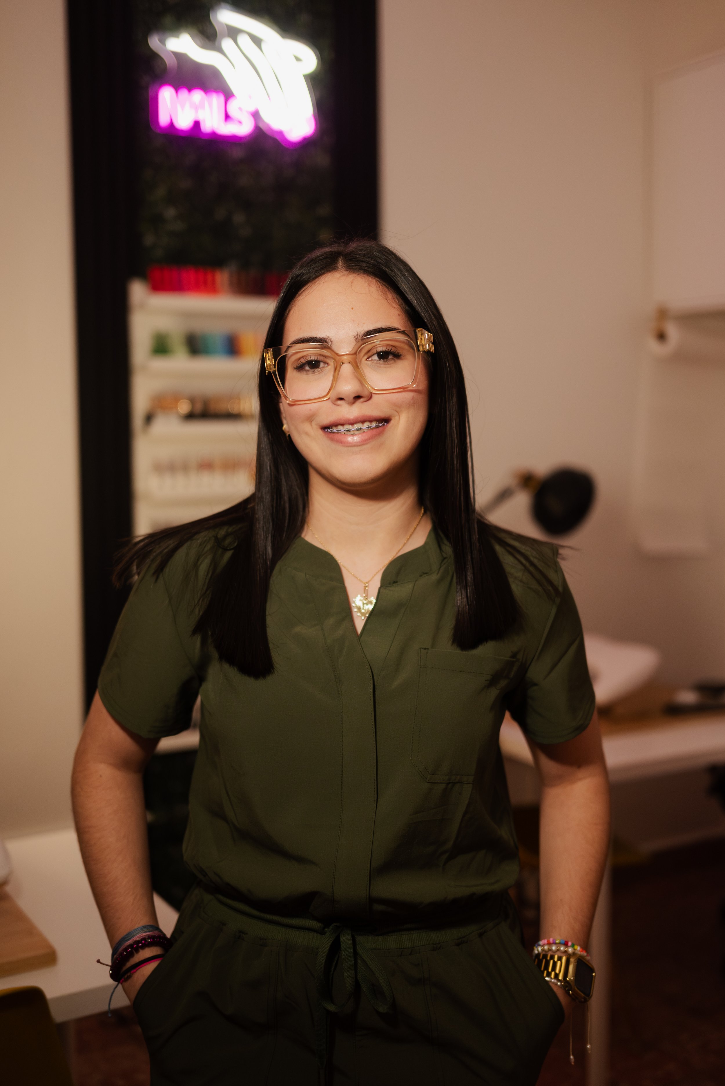 A young woman with straight black hair, glasses, and braces smiling and standing indoors. She is wearing a green scrub top, bracelets, a necklace, and a watch. Behind her is a nail salon sign with neon pink and white lights and a nail polish display.